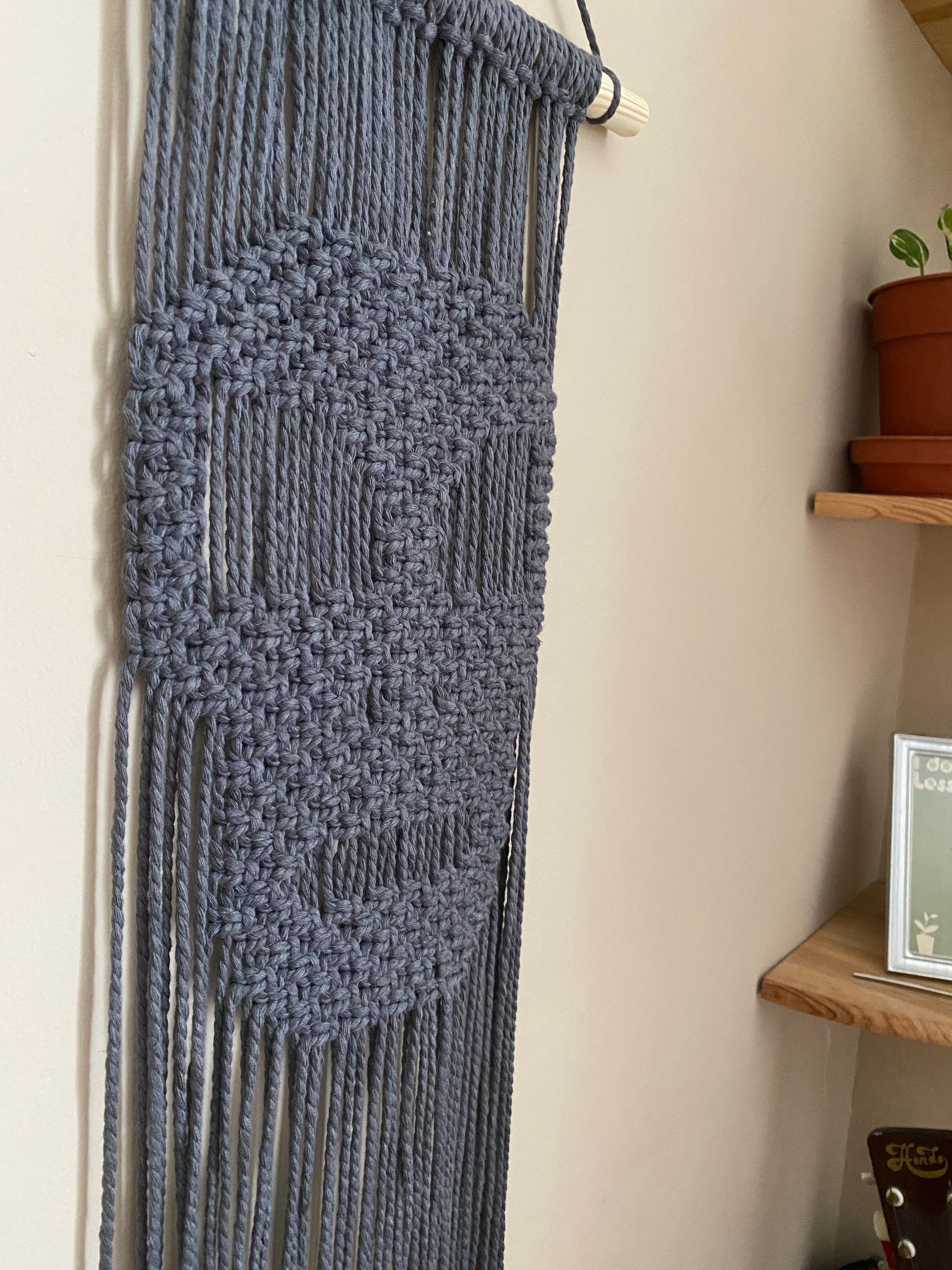Close up of the intricate knots on a dark grey macrame wall hanging with a knotted pattern in the shape of a skull hanging against a beige wall.