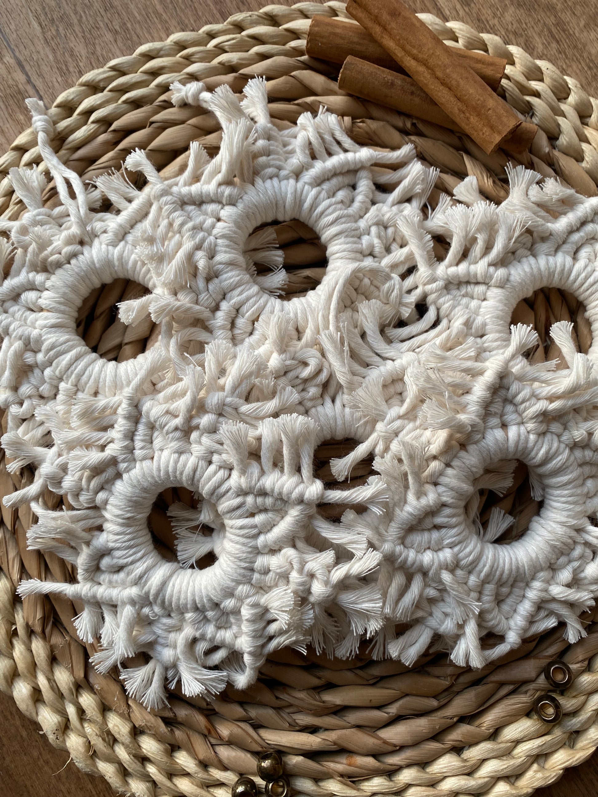 A set of 6 handmade macrame snowflake ornaments, knotted with natural coloured cotton yarn, on a wicker surface surrounded by cinnamon sticks and beads for decoration.