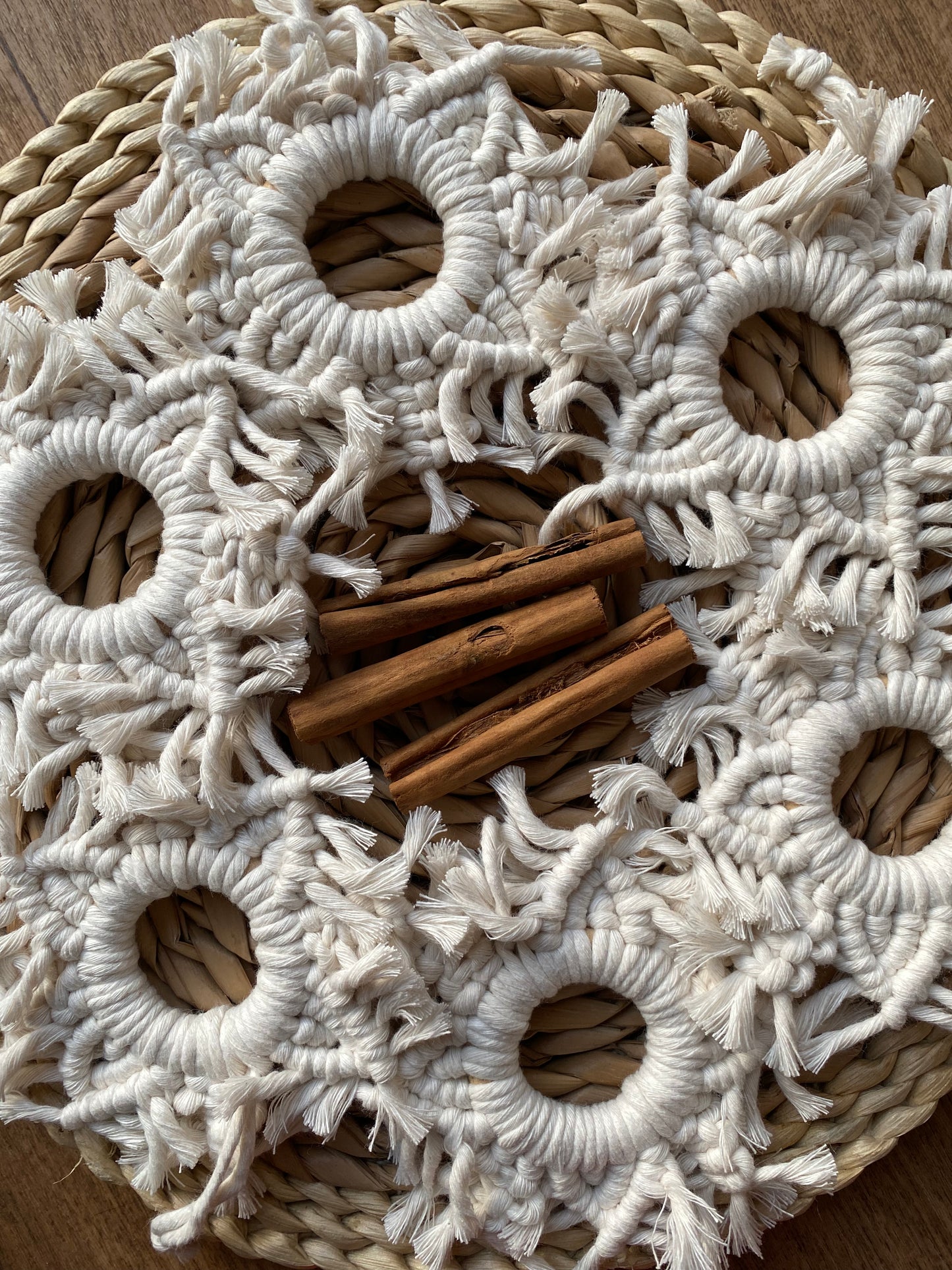 Six handmade macrame snowflake ornaments, knotted with natural coloured cotton yarn, displayed in a circle on a wicker surface with cinnamon sticks in the middle.