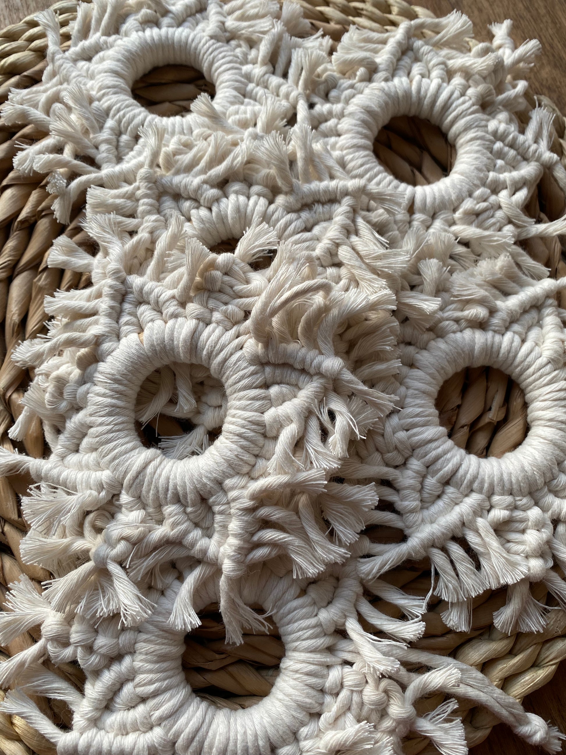 Close up of 6 handmade macrame snowflake ornaments, knotted with natural coloured cotton yarn, piled on top of one another on a wicker surface.