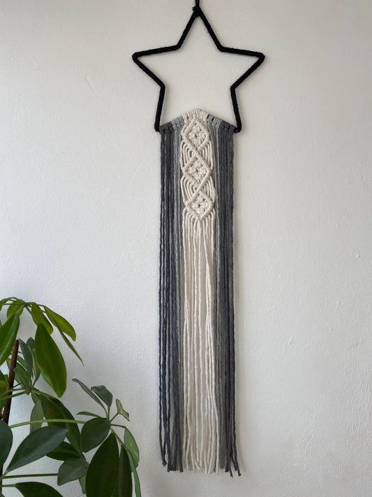 A black star shape wall hanging with long monochrome hanging tails, hanging on a white wall, next to a green houseplant.