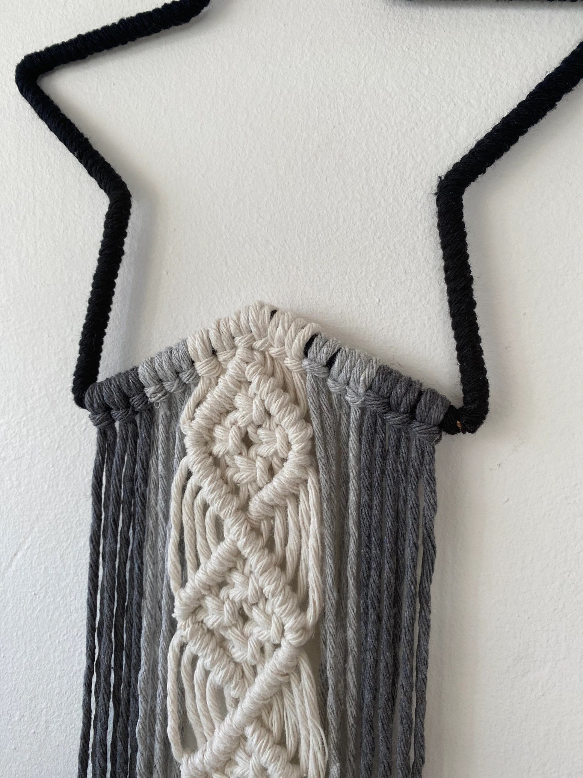 A close up image of the knotted section on a macrame wall hanging with long monochrome hanging tails, hanging on a white wall.