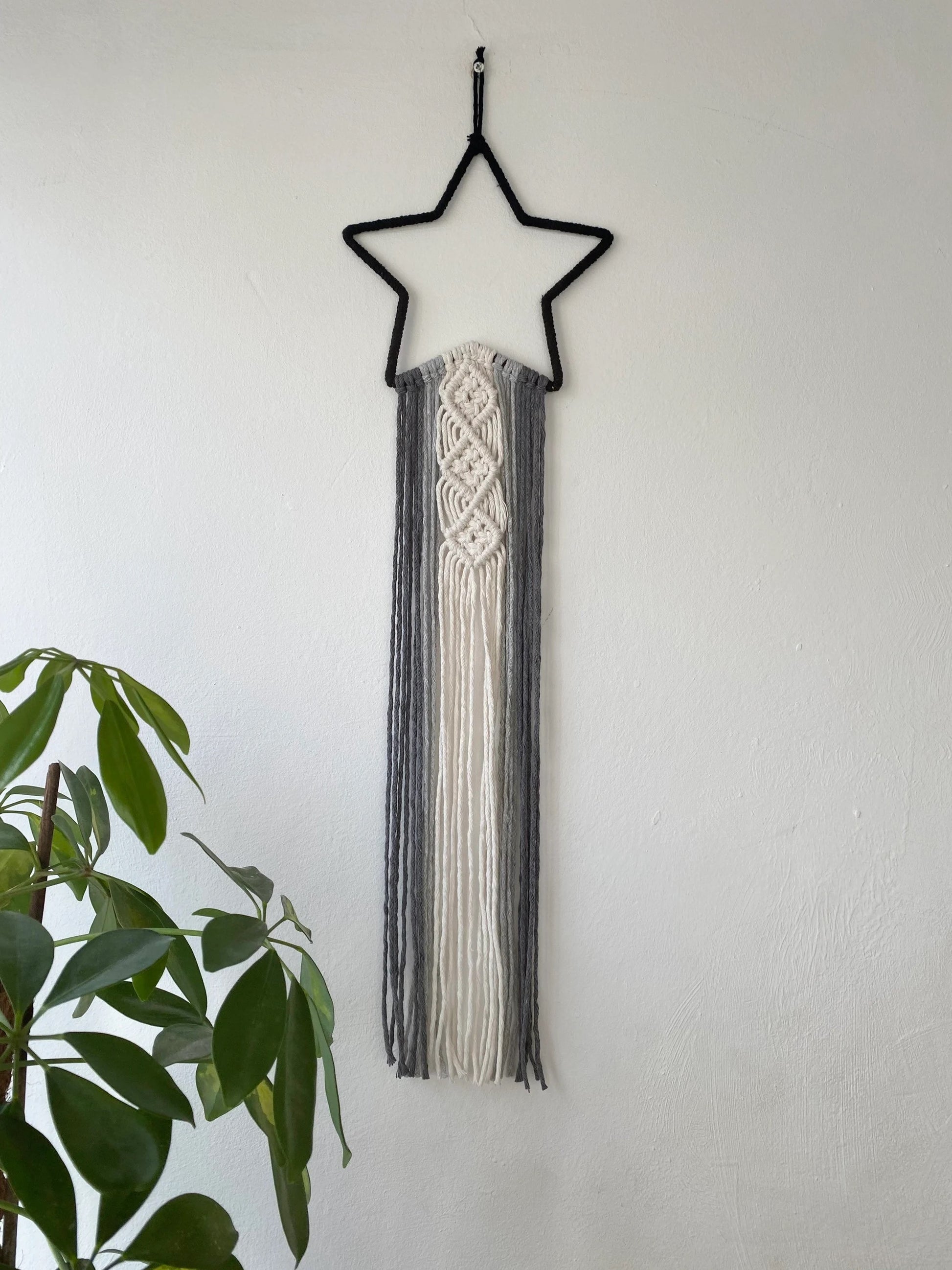 A black star shape wall hanging with long monochrome hanging tails, hanging on a white wall, next to a green houseplant.
