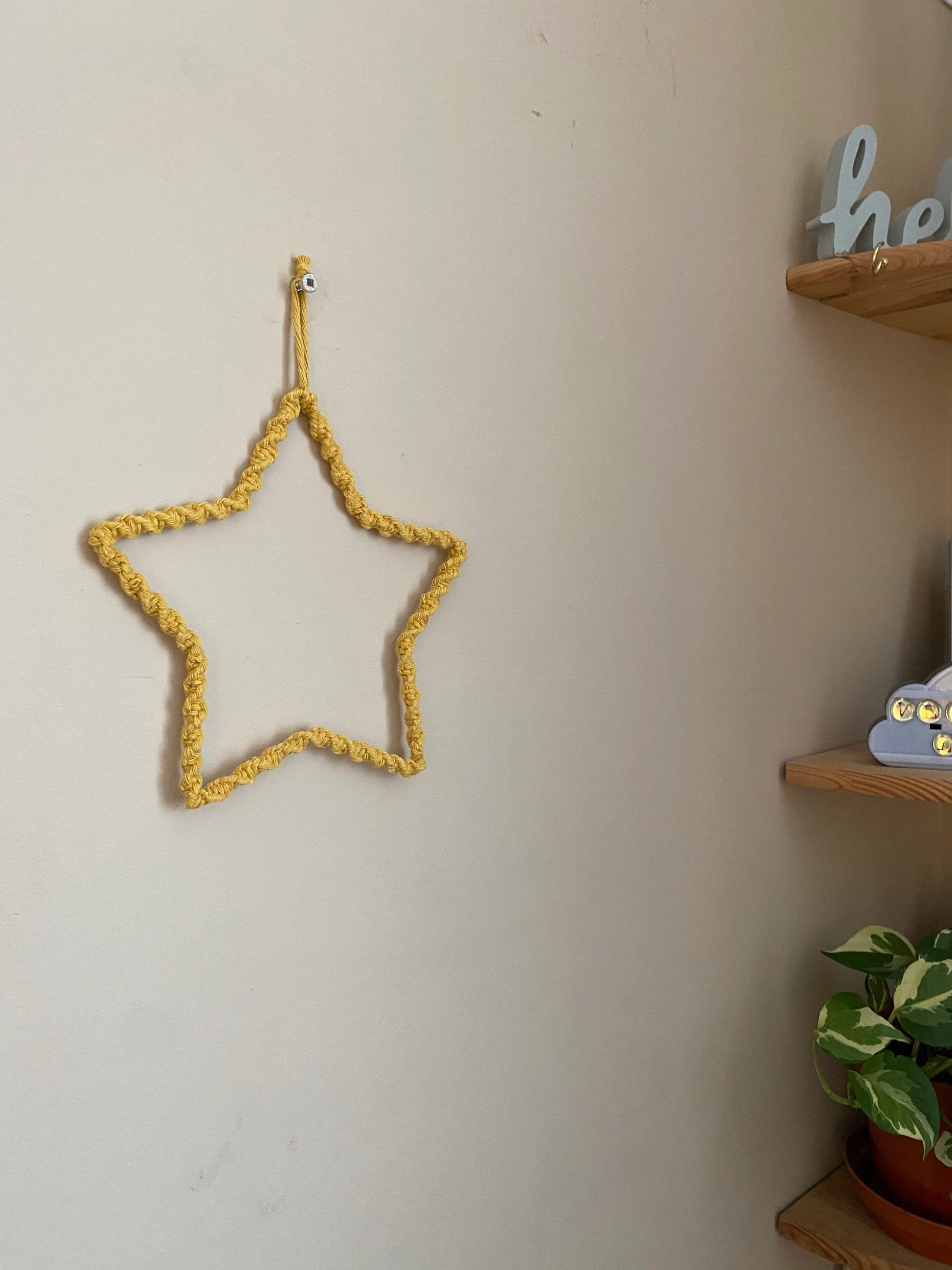 Star shaped wall hanging, wrapped in mustard yellow cotton with a twisted knot pattern, hanging on a beige wall. There is a set of wooden corner shelves to the right.