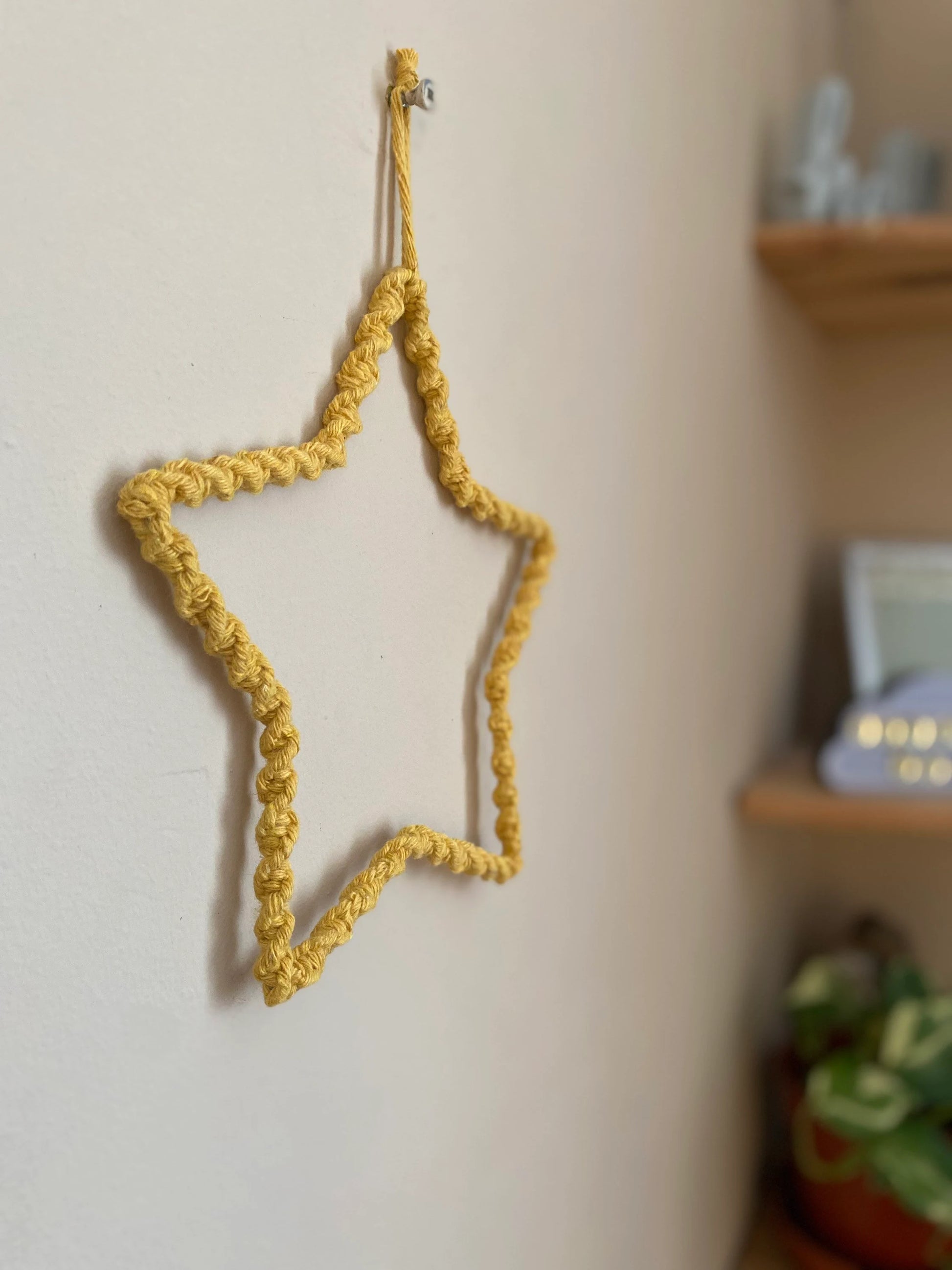 Star shaped wall hanging, wrapped in mustard yellow cotton with a twisted knot pattern, hanging on a beige wall. There is a set of wooden corner shelves just out of focus to the right.
