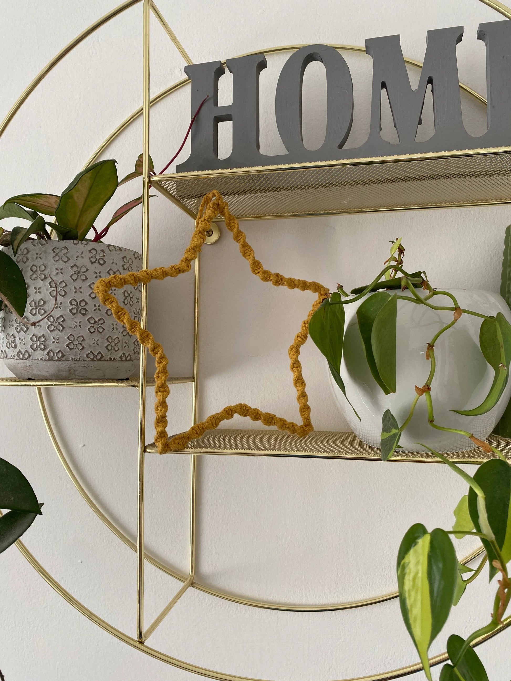 Star shaped decoration, wrapped in mustard yellow cotton with a twisted knot pattern, placed on a gold metal circular shelf, in between two houseplants. There is also a grey wooden sign that says HELLO on the shelf above.