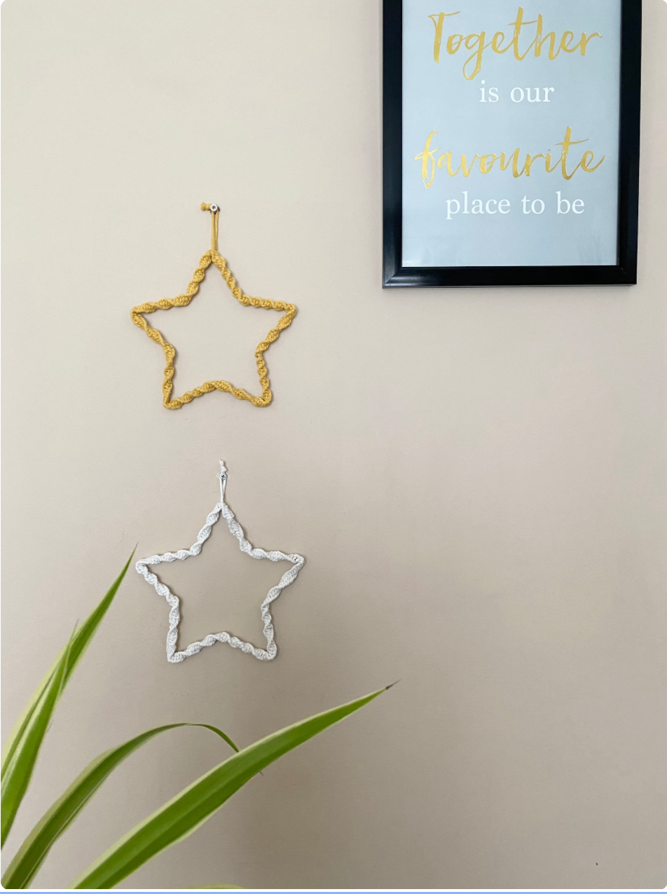 Two star shaped wall hangings, wrapped in cotton with a twisted knot pattern, hanging one above the other on a beige wall. The top star is mustard yellow and the bottom star is natural coloured. There is a green houseplant visible at the bottom of the image and a framed print to the top right of the image.