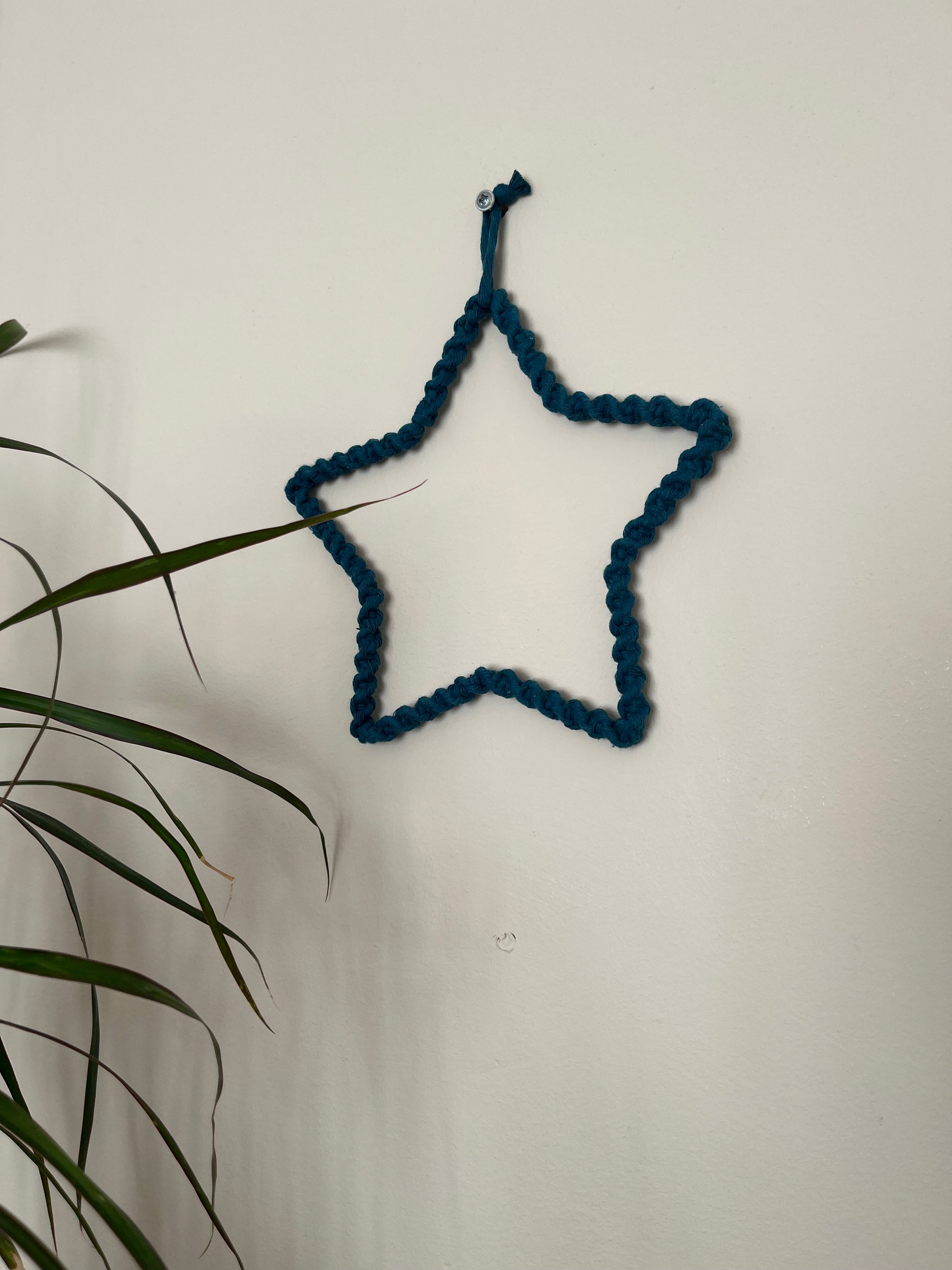 Star shaped wall hanging, wrapped in petrol blue cotton with a twisted knot pattern, hanging on a beige wall. There is a green houseplant in the bottom left of the image.
