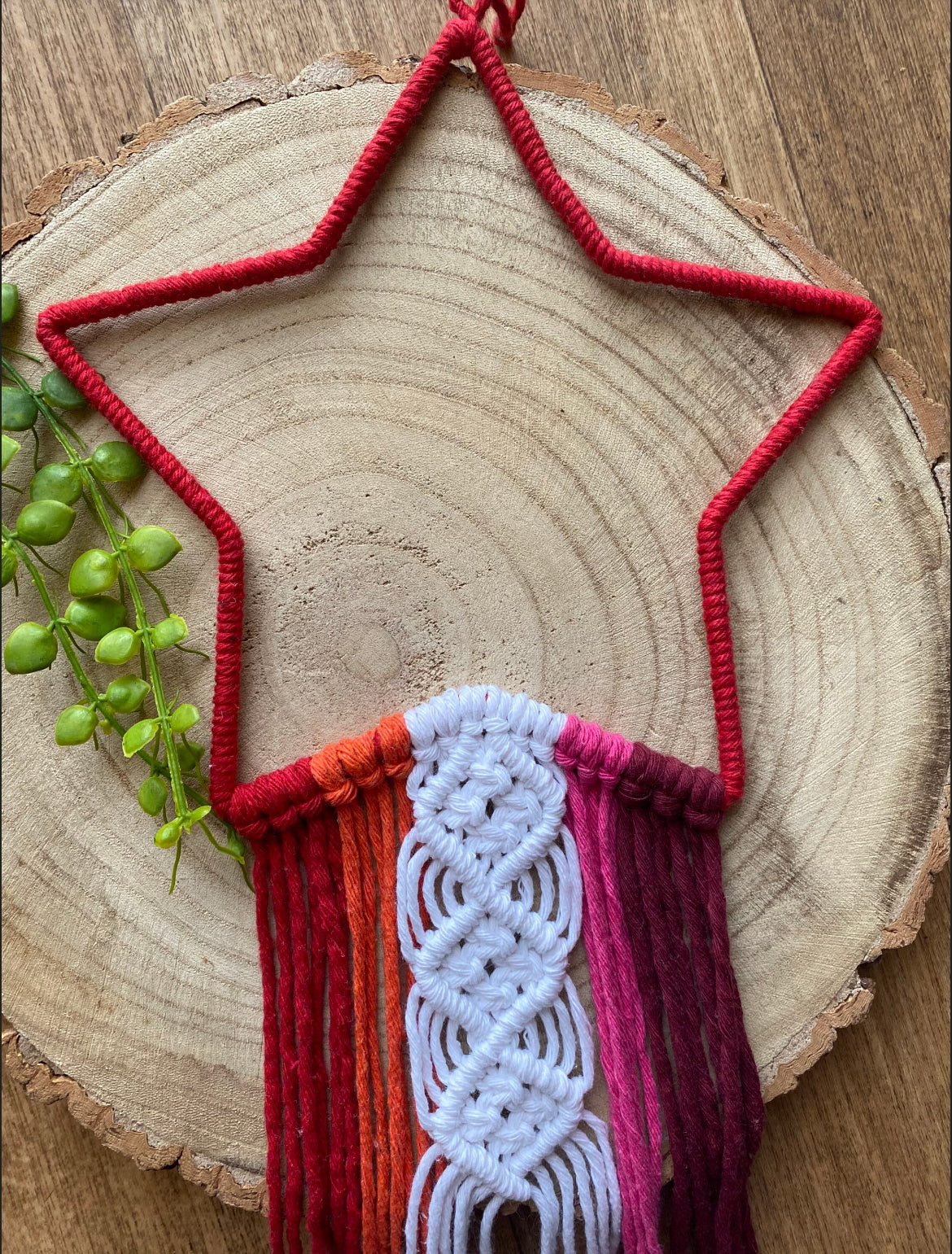 A close up of a pink macrame star shape wall hanging with red, orange and pink hanging tails, laid on a wooden board.