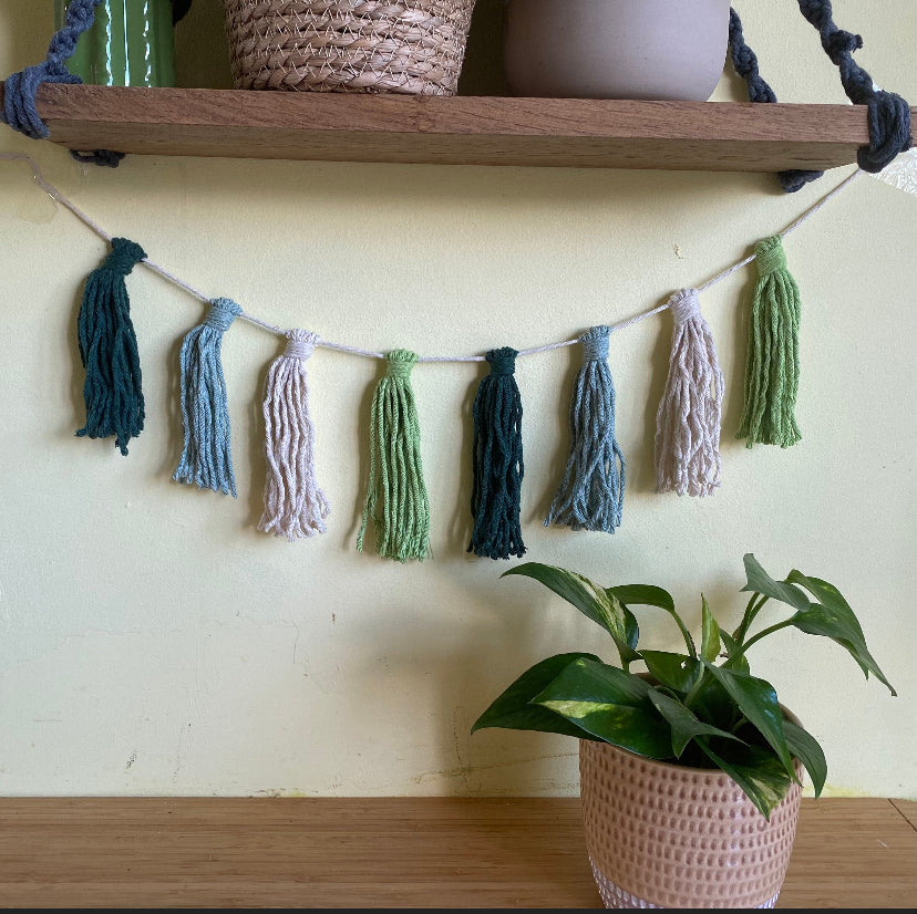 Macrame tassel garland, in shades of green, hanging on a wall underneath a wooden shelf. There is a small green plant on a table nearby.