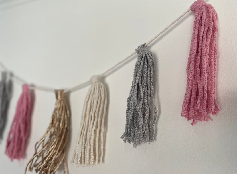 Close up of a macrame tassel garland, in pink, grey, natural and metallic yarns, hanging on a white wall.