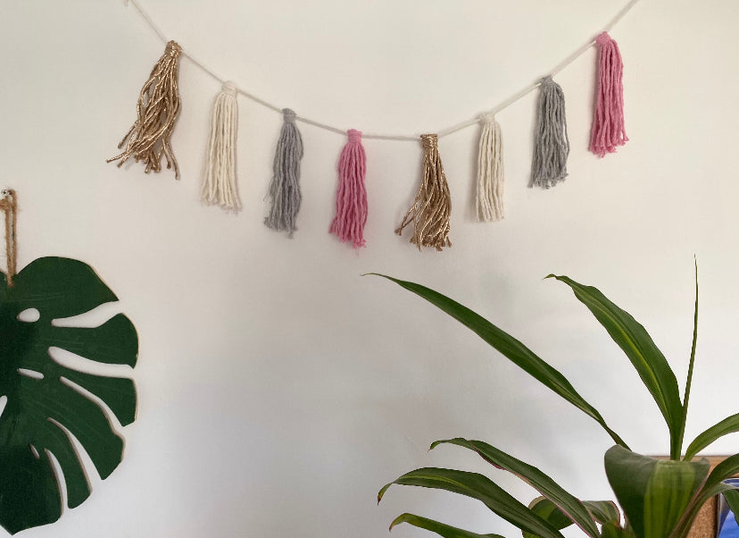 Macrame tassel garland, in pink, grey, natural and metallic yarn, hanging on a white wall. There is a green houseplant nearby, and a large green wooden leaf hanging on the wall.
