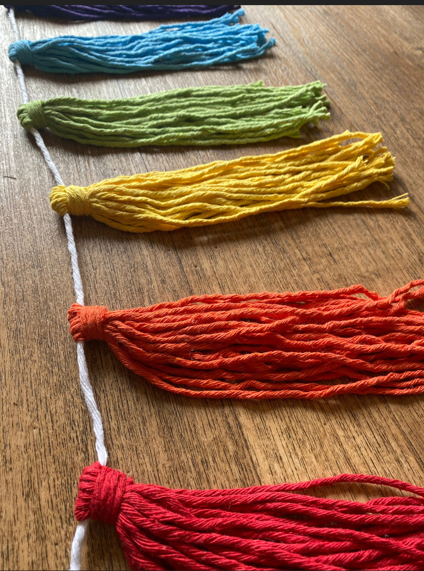 Close up of a macrame tassel garland, in rainbow colours laid out in a line on a wooden floor.