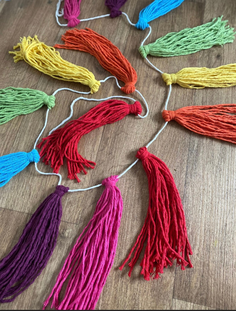 Close up of a macrame tassel garland, in rainbow colours, laid out on a wooden floor.
