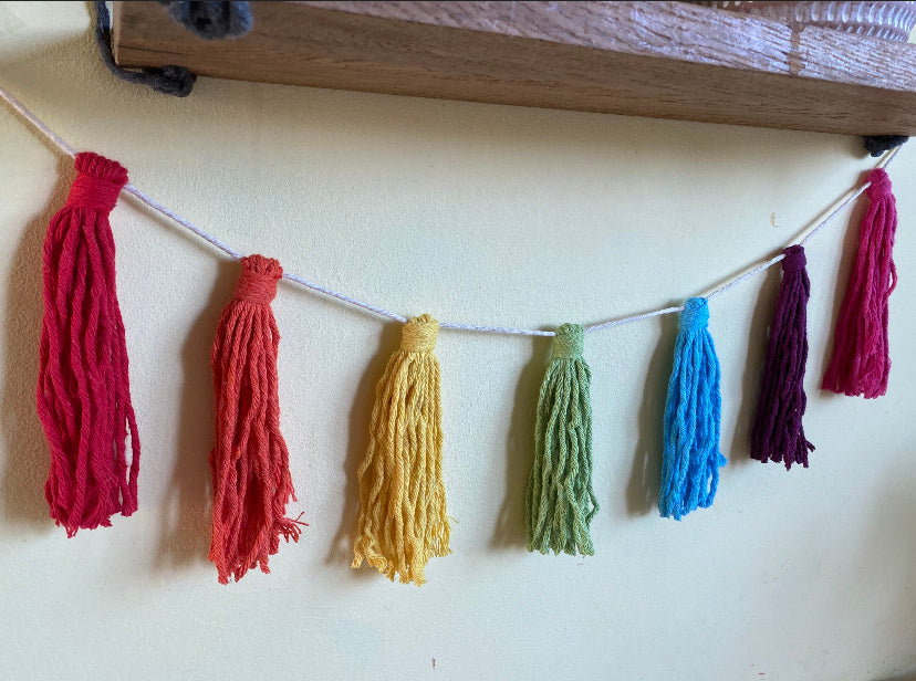 Macrame tassel garland, in rainbow colours, hanging on a wall underneath a wooden shelf.