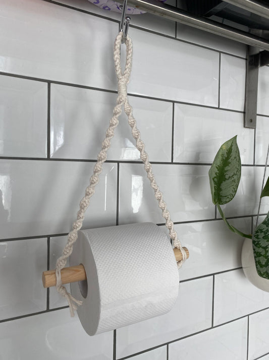 A white toilet roll is held on a handmade macrame toilet roll holder, made with natural recycled cotton and a wooden dowel.