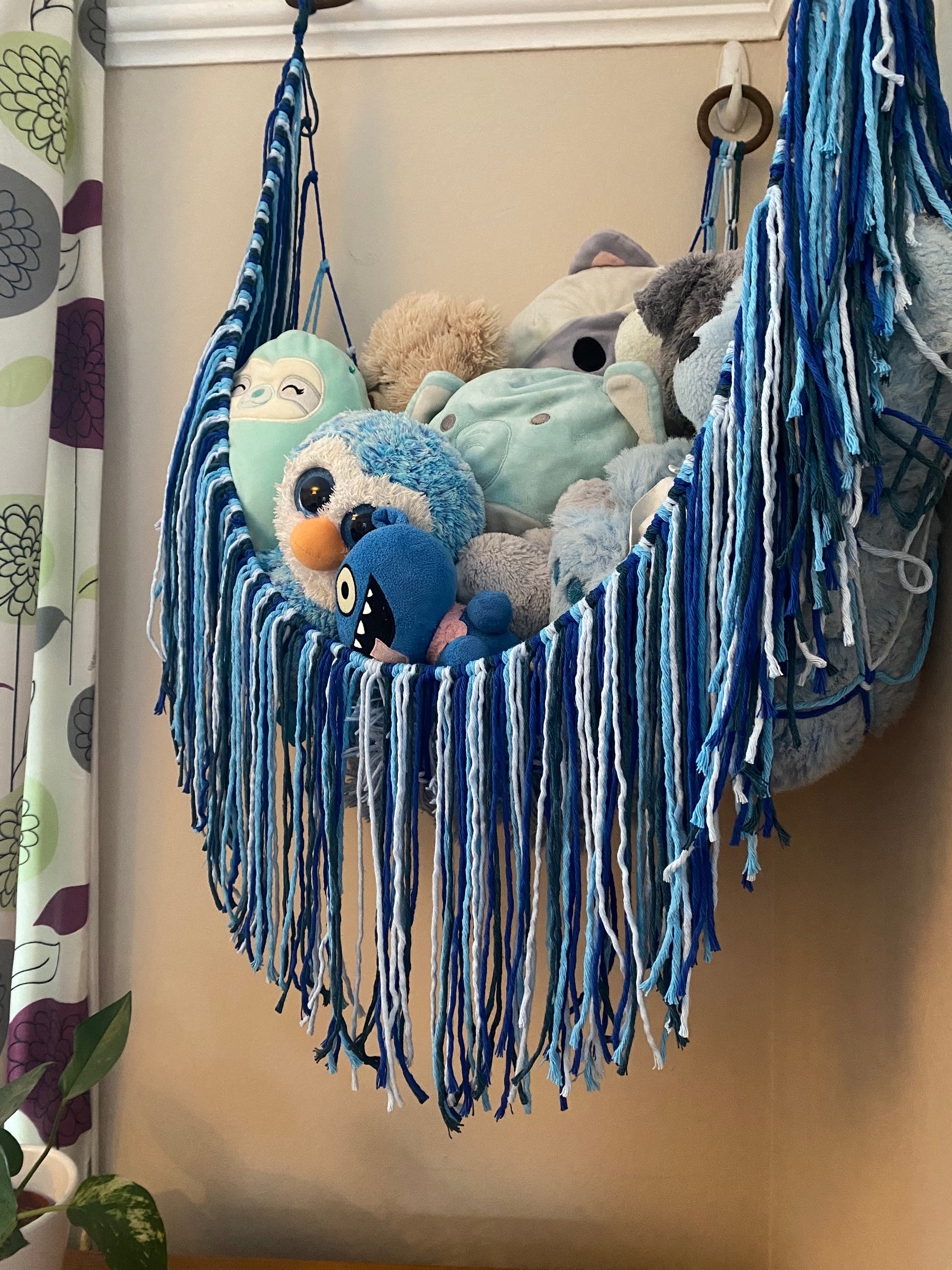 Close up of a macrame toy hammock made with blue coloured yarn, with a long hanging fringe, filled with soft toys.