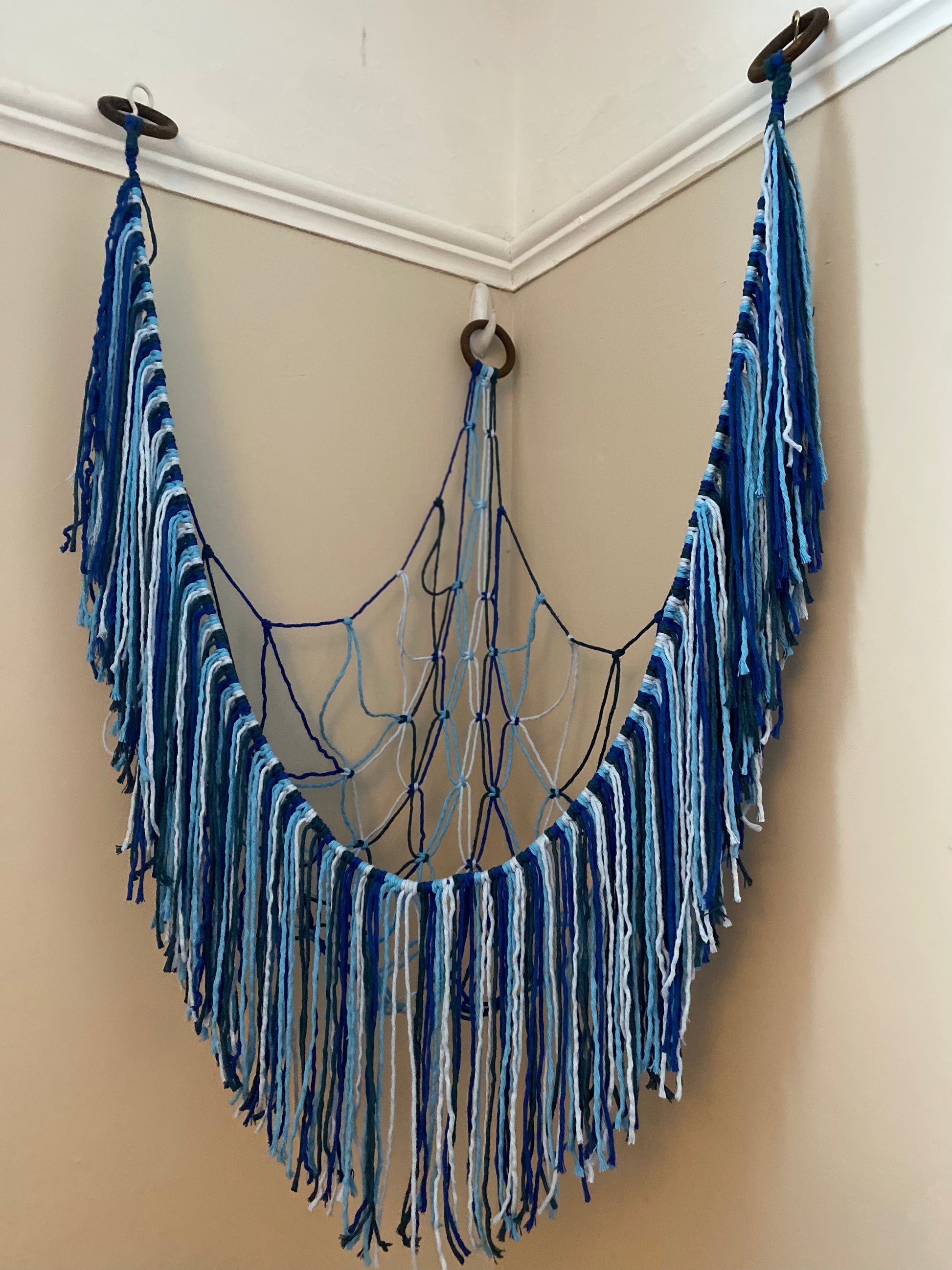 An empty macrame toy hammock made with blue yarn, with a long hanging fringe, hangs in the corner of a room with beige painted walls.