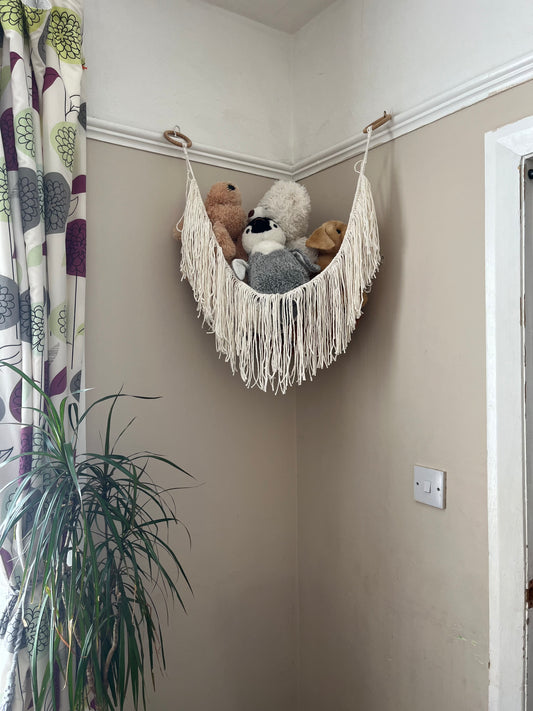 A macrame toy hammock made with natural coloured yarn, with a long hanging fringe, hangs in the corner of a room with beige painted walls, filled with soft toys.