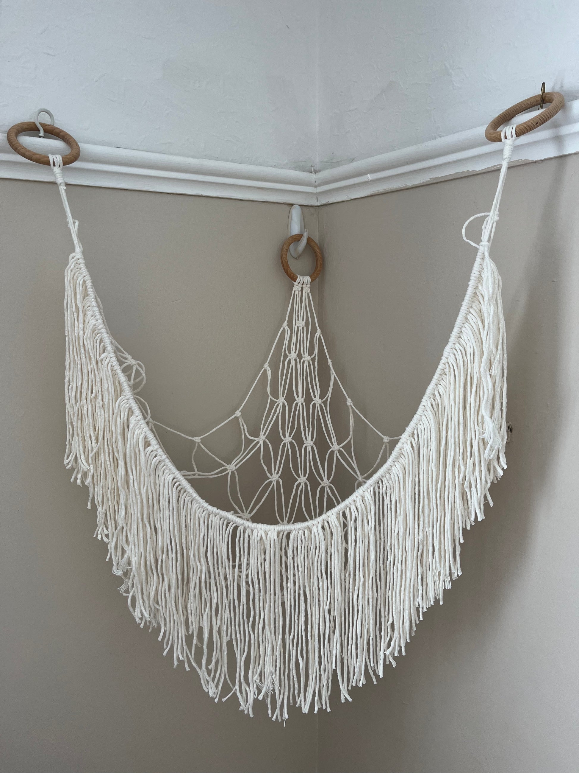 An empty macrame toy hammock made with natural coloured yarn, with a long hanging fringe, hangs in the corner of a room with beige painted walls.