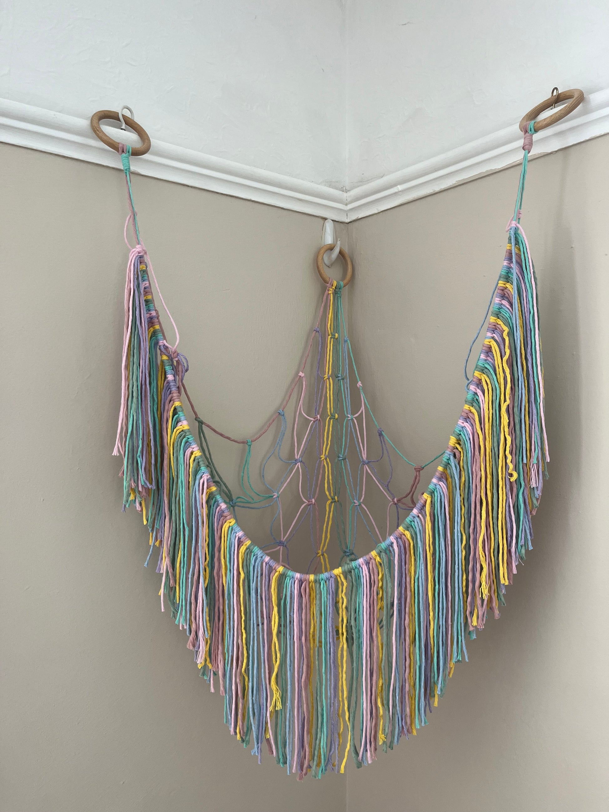 An empty macrame toy hammock made with pastel coloured yarn, with a long hanging fringe, hangs in the corner of a room with beige painted walls.