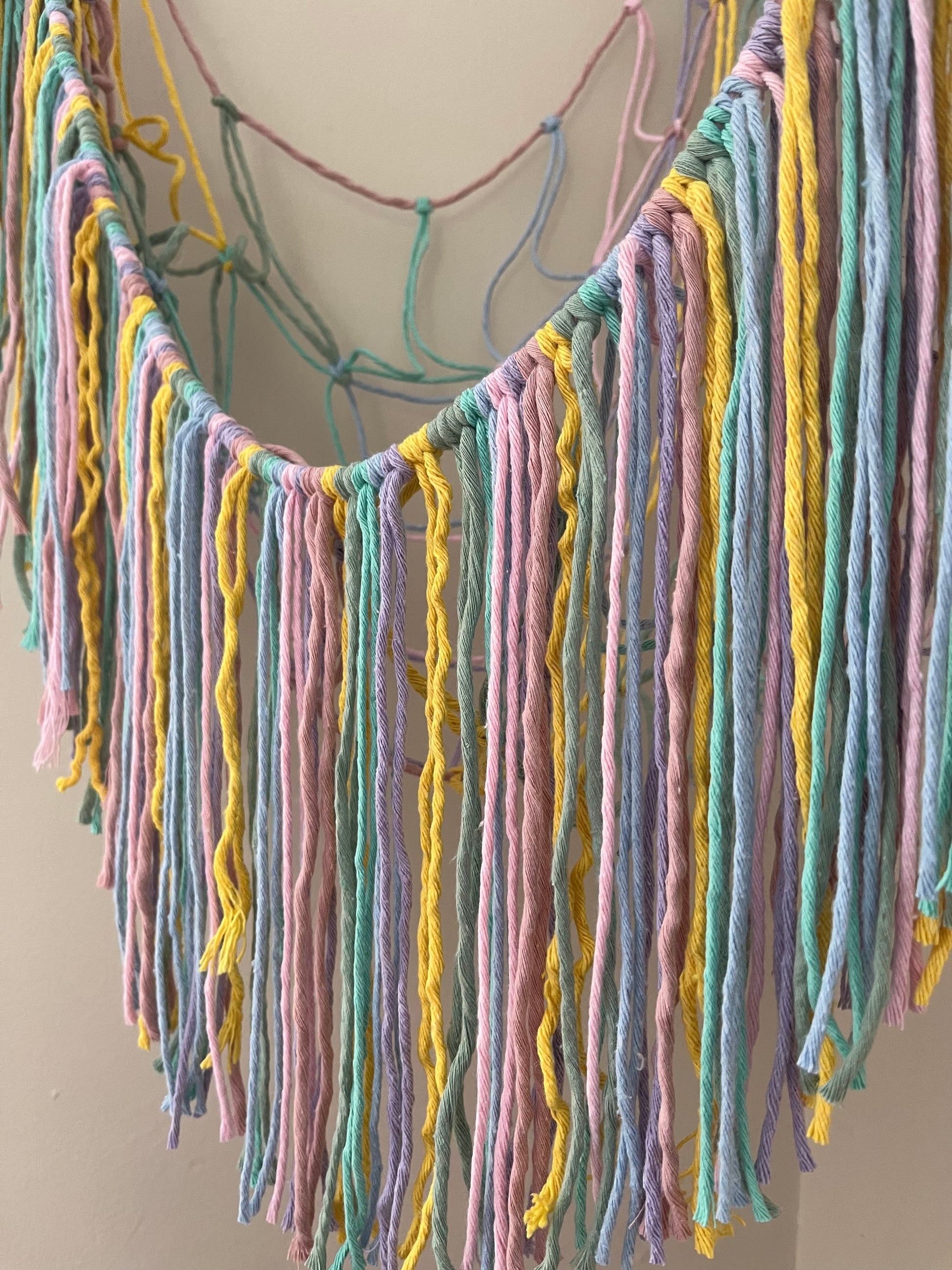 A close up of the long hanging fringe, on a macrame toy hammock, made with pastel coloured yarn.