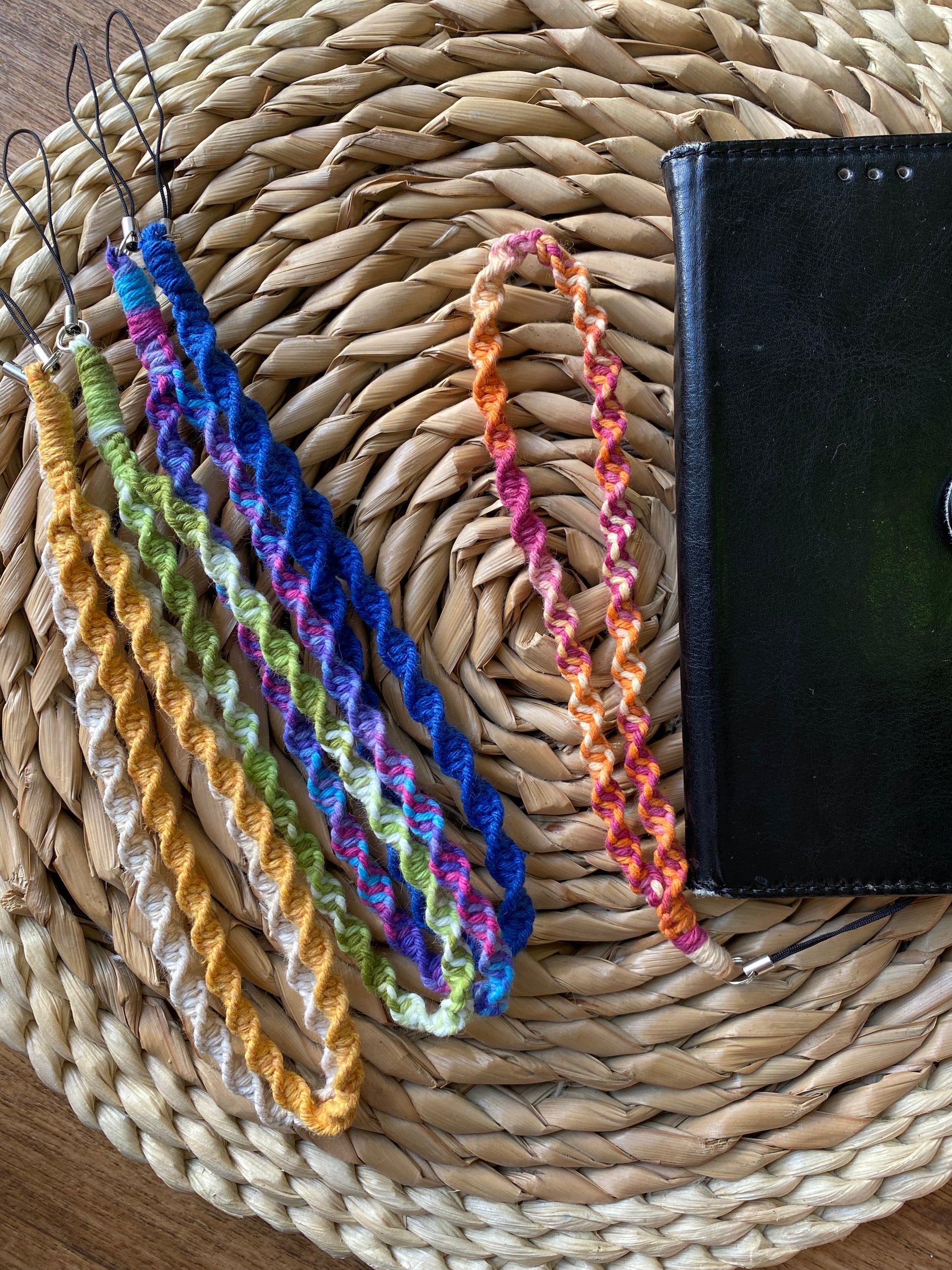 A bundle of handmade macrame phone charms, in a variety of colours lay on a woven placemat. There is also a black phone case on the mat with a further phone charm attached to the bottom of it.