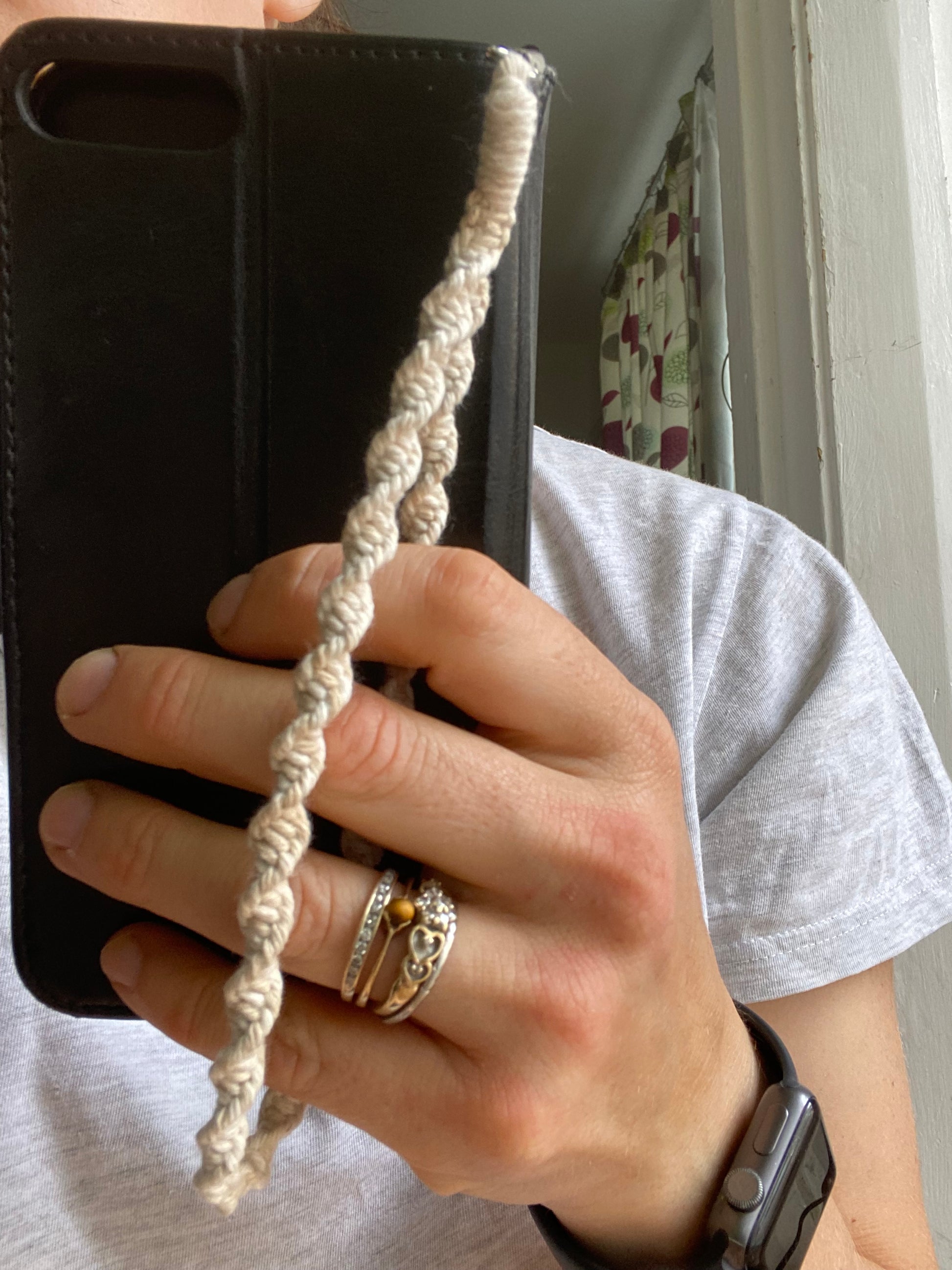 A woman holds a black wallet style phone case, which has a beige macrame phone wristlet charm attached to it.