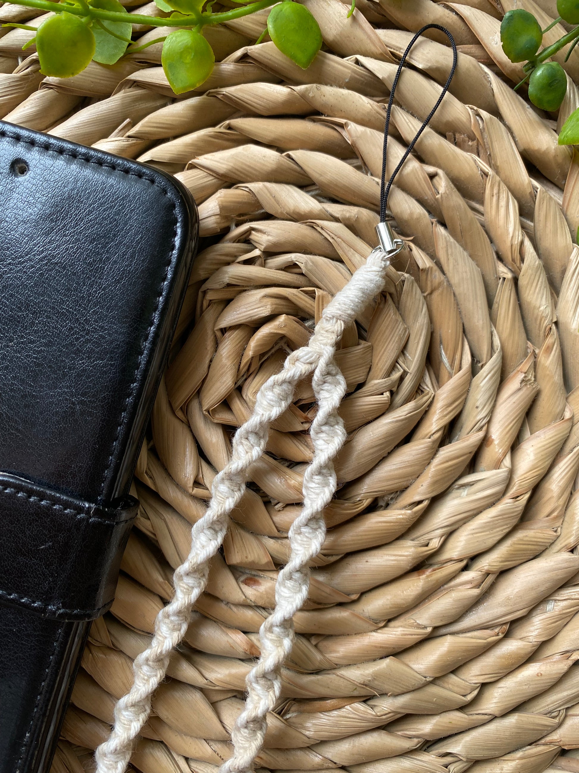 A handmade macrame phone charm made with beige coloured yarn lays on a wicker placemat beside a black wallet style phone case.