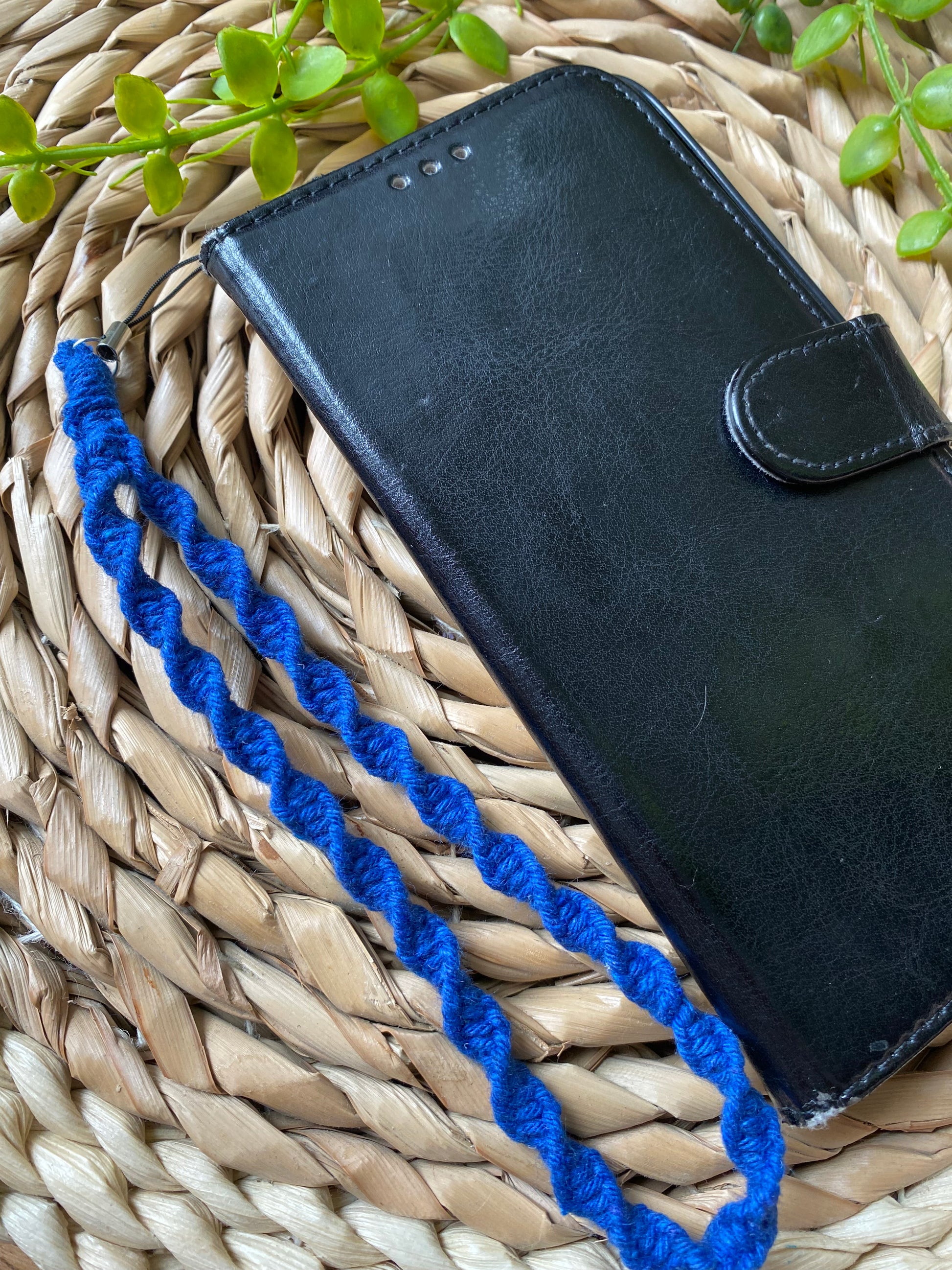 A black wallet style phone case lays on a woven wicker placemat. The phone case has a macrame phone charm made with blue coloured yarn attached to it at the top.