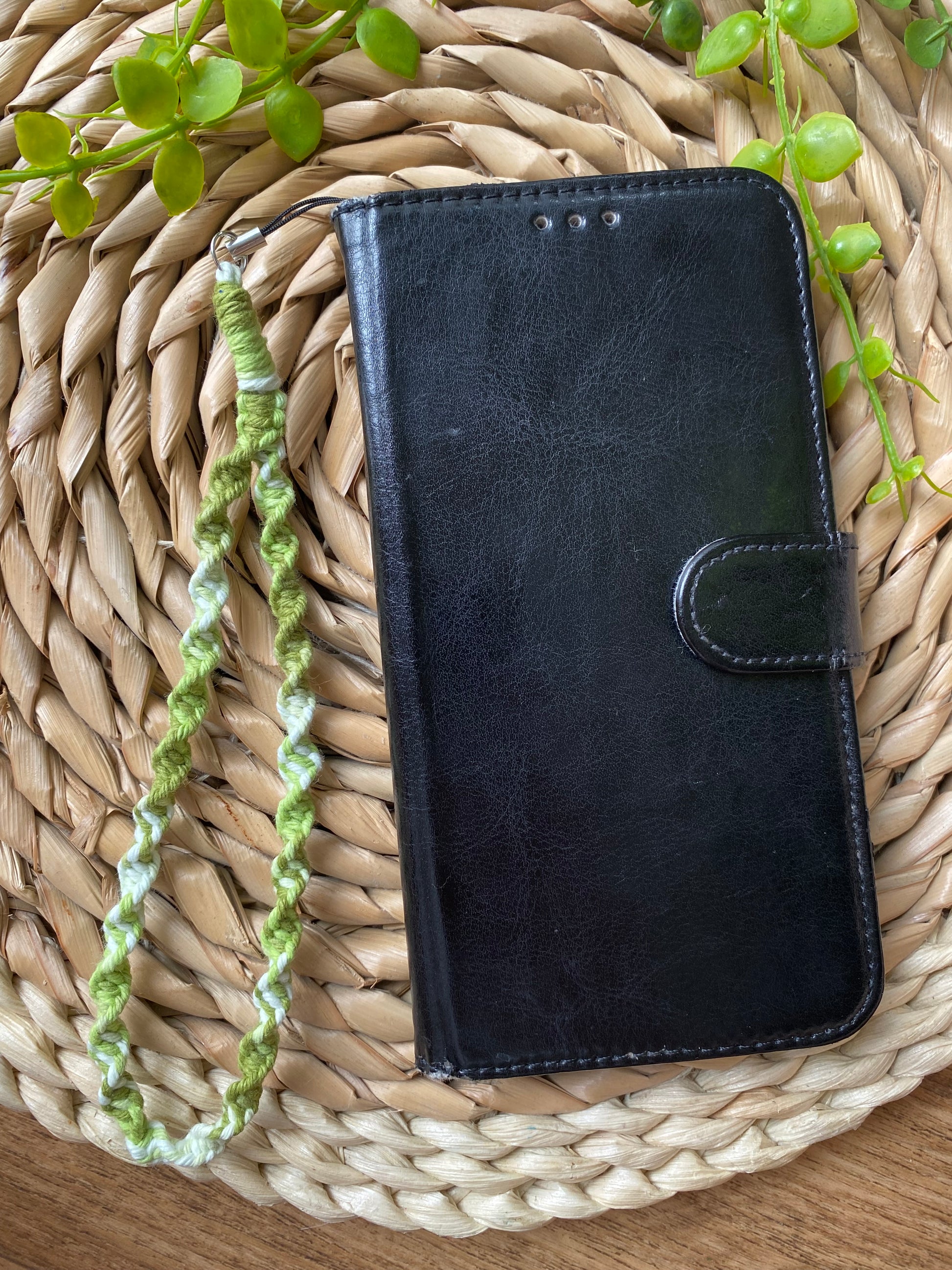 A black wallet style phone case lays on a woven wicker placemat. The phone case has a macrame phone charm made with green coloured yarn attached to it at the top.