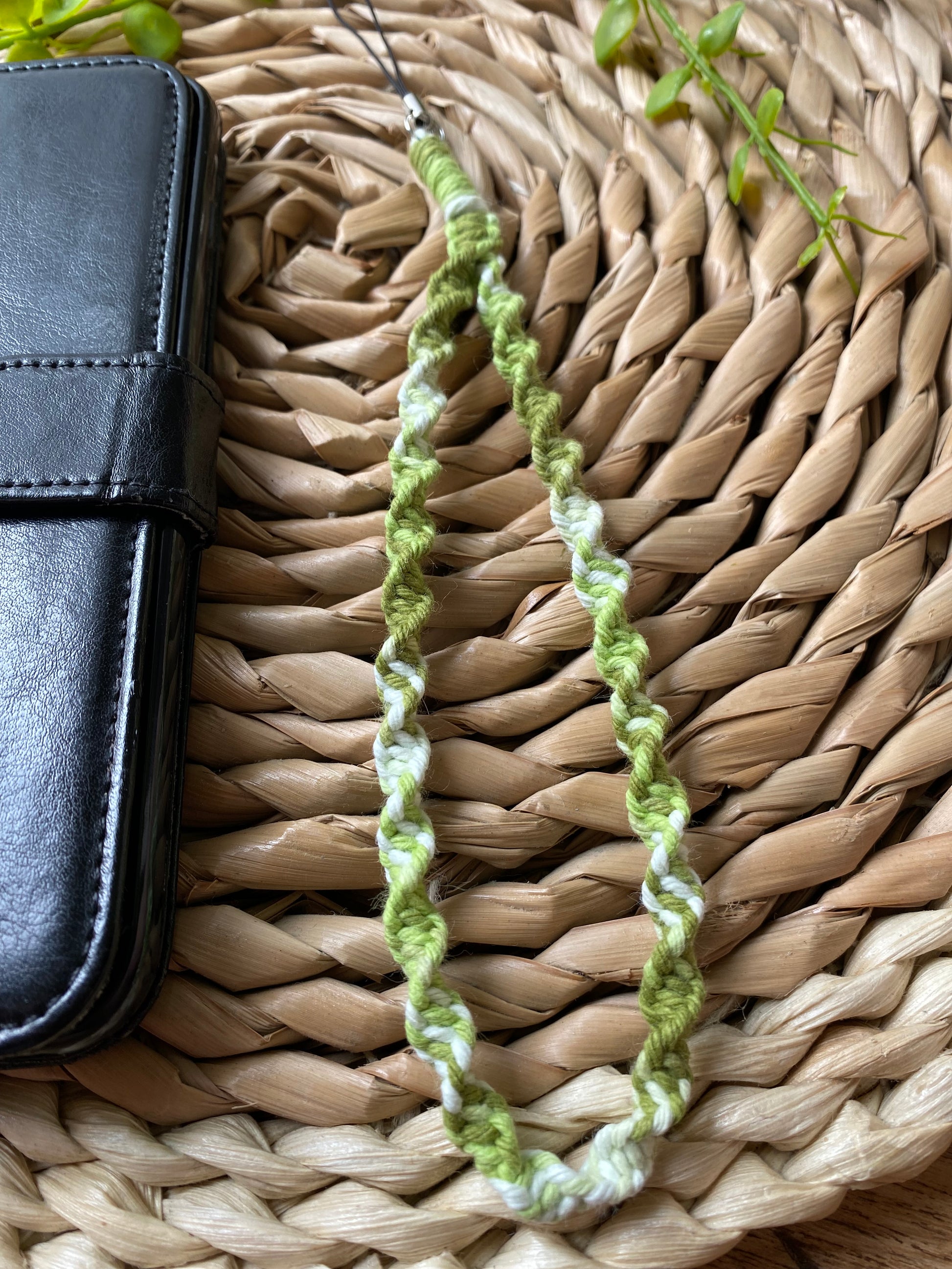 A macrame phone charm made with green coloured yarn lays on a woven wicker placemat beside a black wallet style phone case.