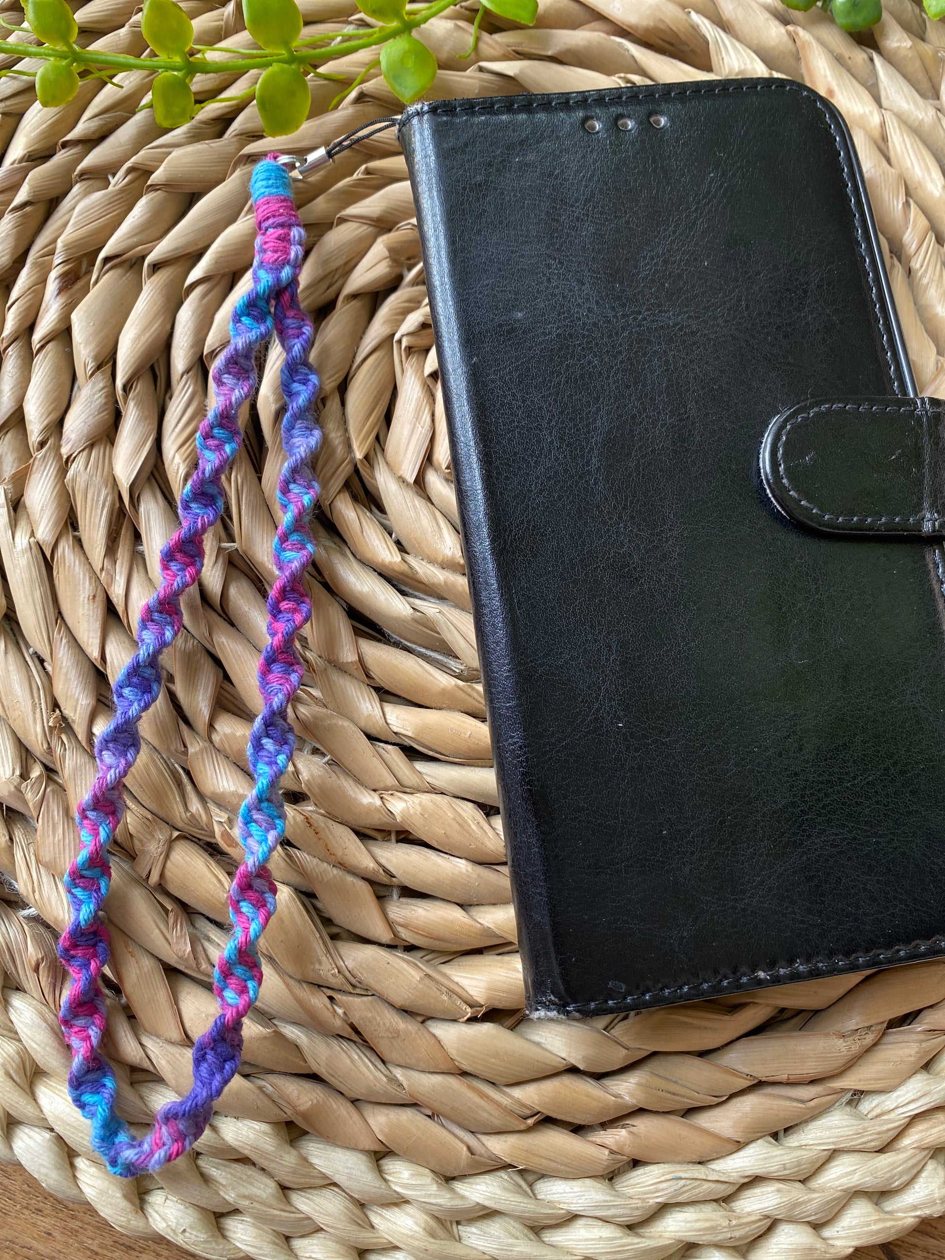 A black wallet style phone case lays on a woven wicker placemat. The phone case has a macrame phone charm made with purple coloured yarn attached to it at the top.