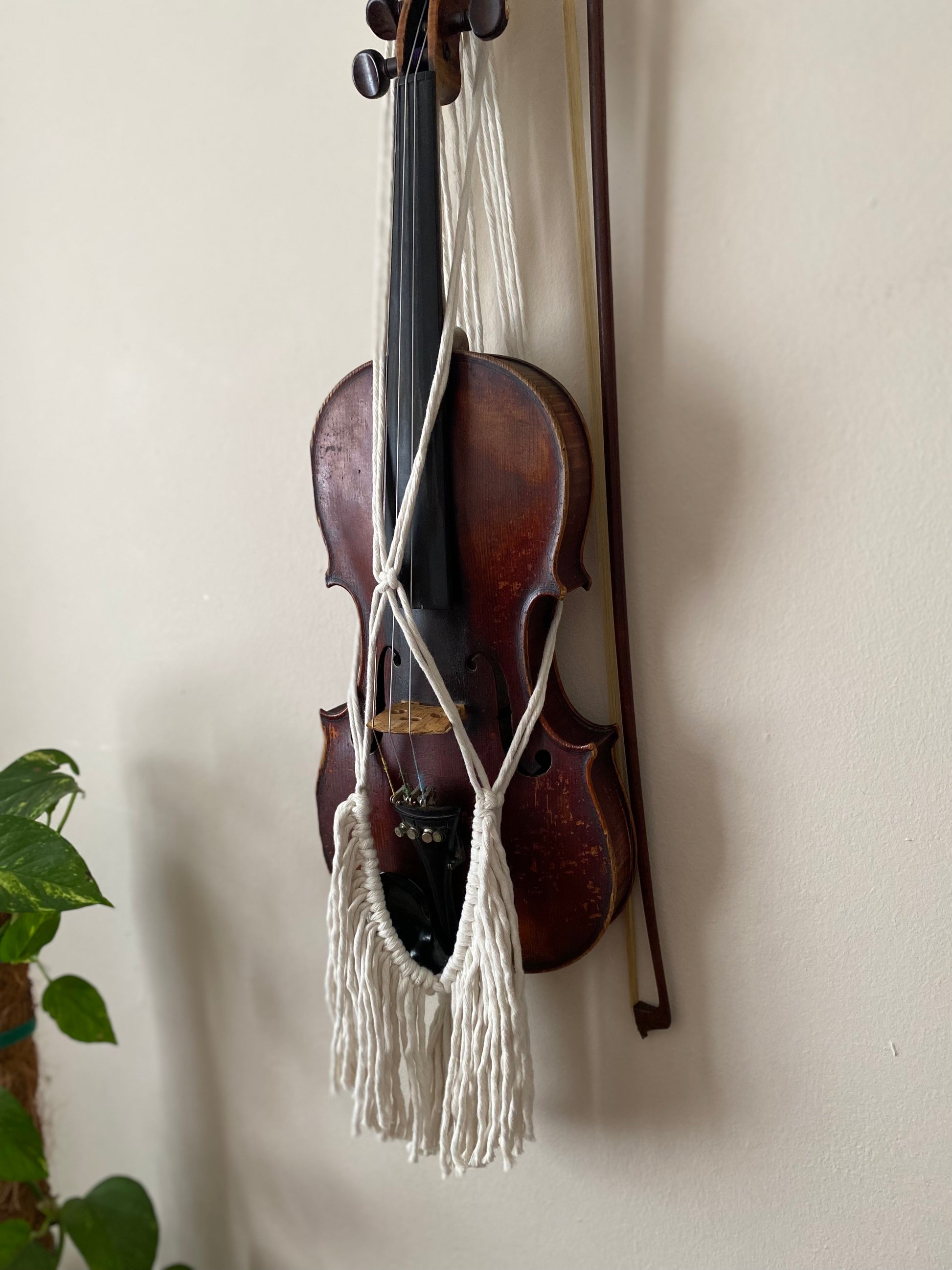 A dark wooden violin hanging in a macrame wall hanger, made with natural off white coloured recycled cotton with boho tassel fringe.