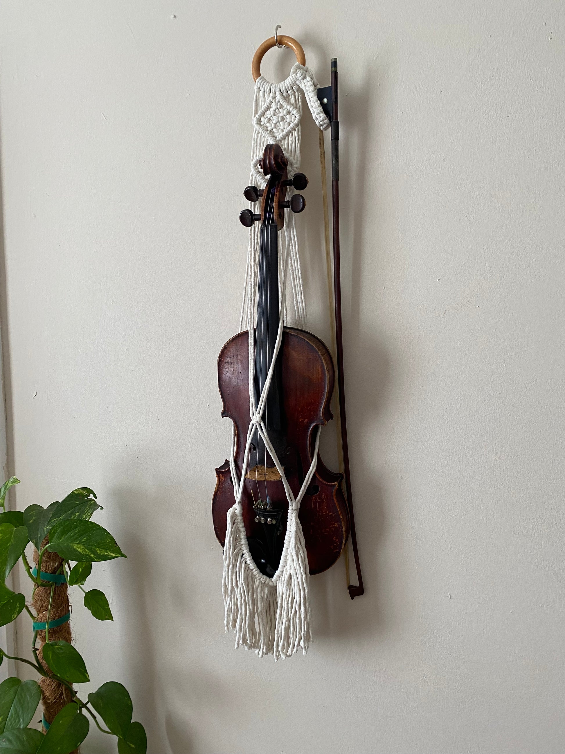 A dark wooden violin hanging in a macrame wall hanger, made with natural off white coloured recycled cotton with long fringe on a wooden ring.