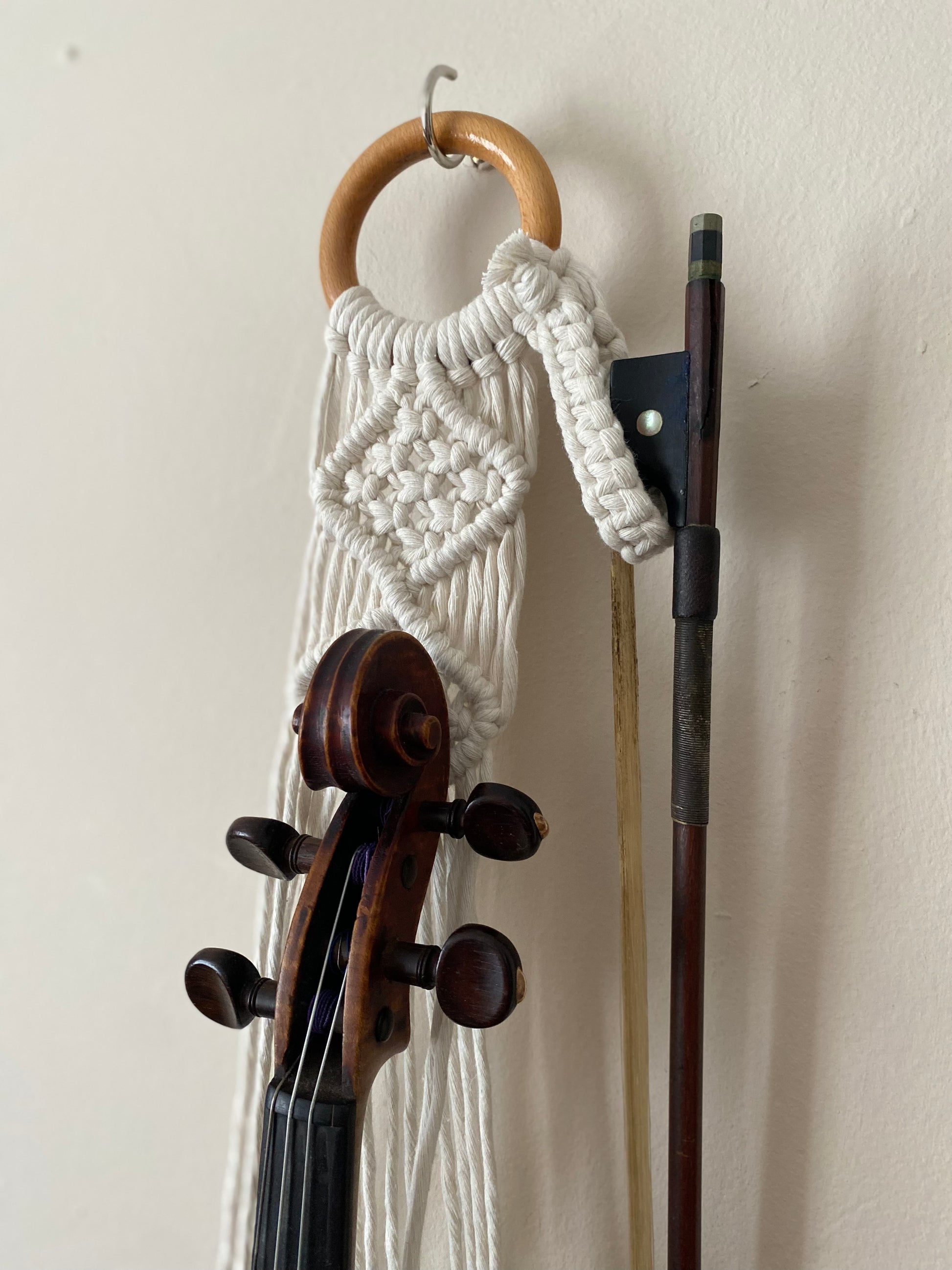 A dark wooden violin and bow hanging in a macrame wall hanger, made with natural off white coloured recycled cotton on a wooden ring.