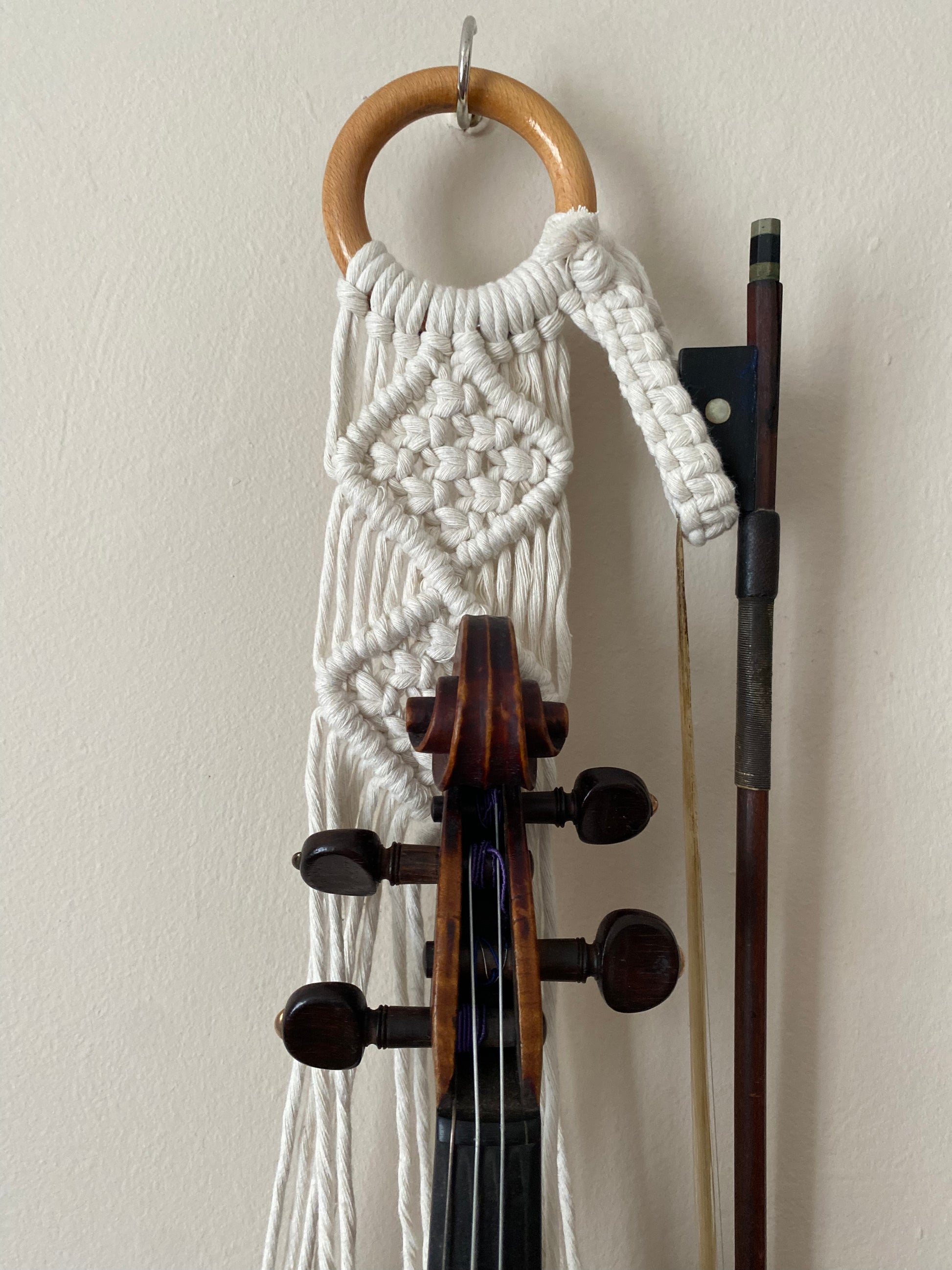 A dark wooden violin and bow hanging in a macrame wall hanger, made with natural off white coloured recycled cotton on a wooden ring.
