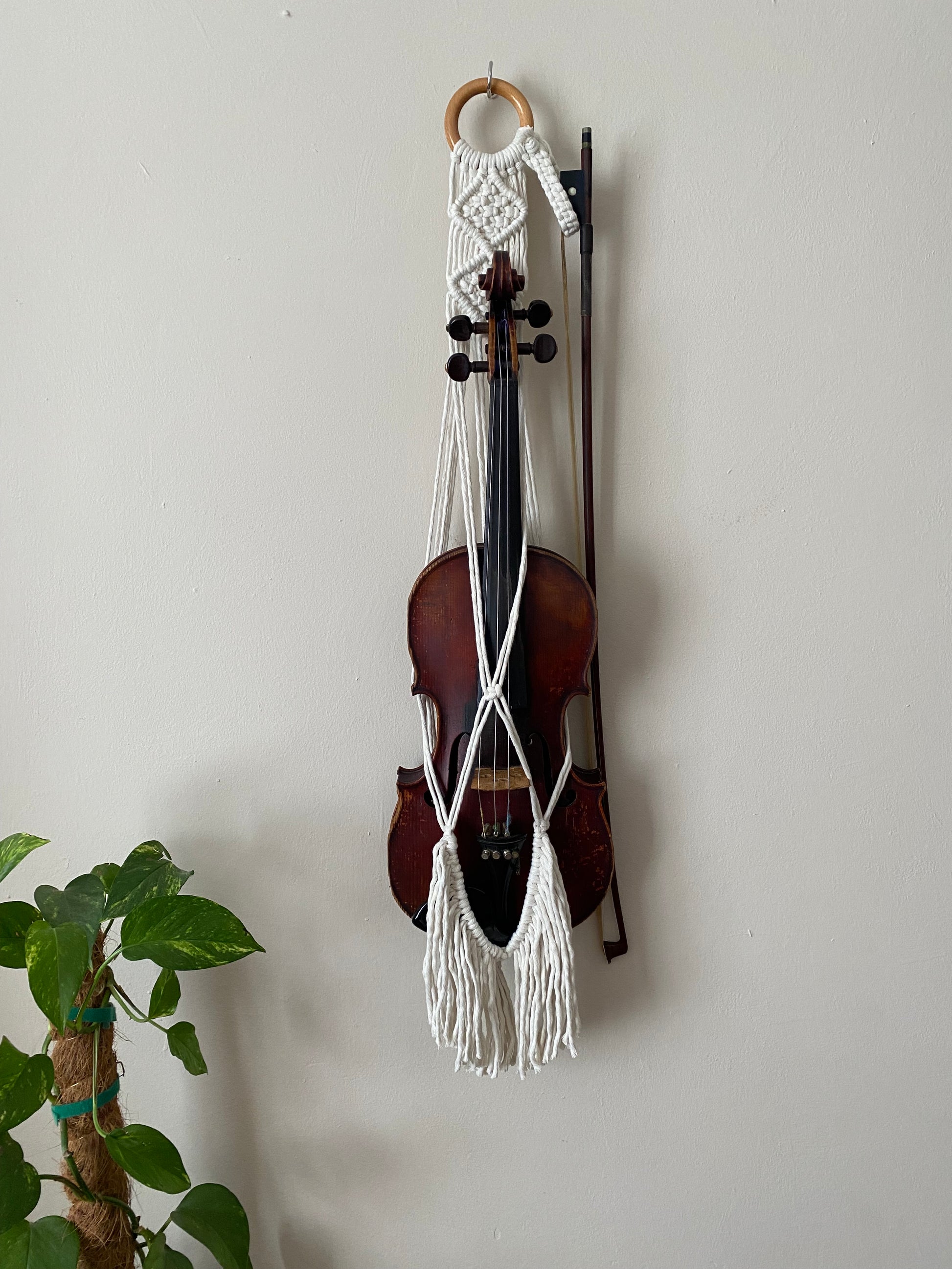 A dark wooden violin and bow hanging in a macrame wall hanger, made with natural off white coloured recycled cotton on a wooden ring, with long boho fringe.