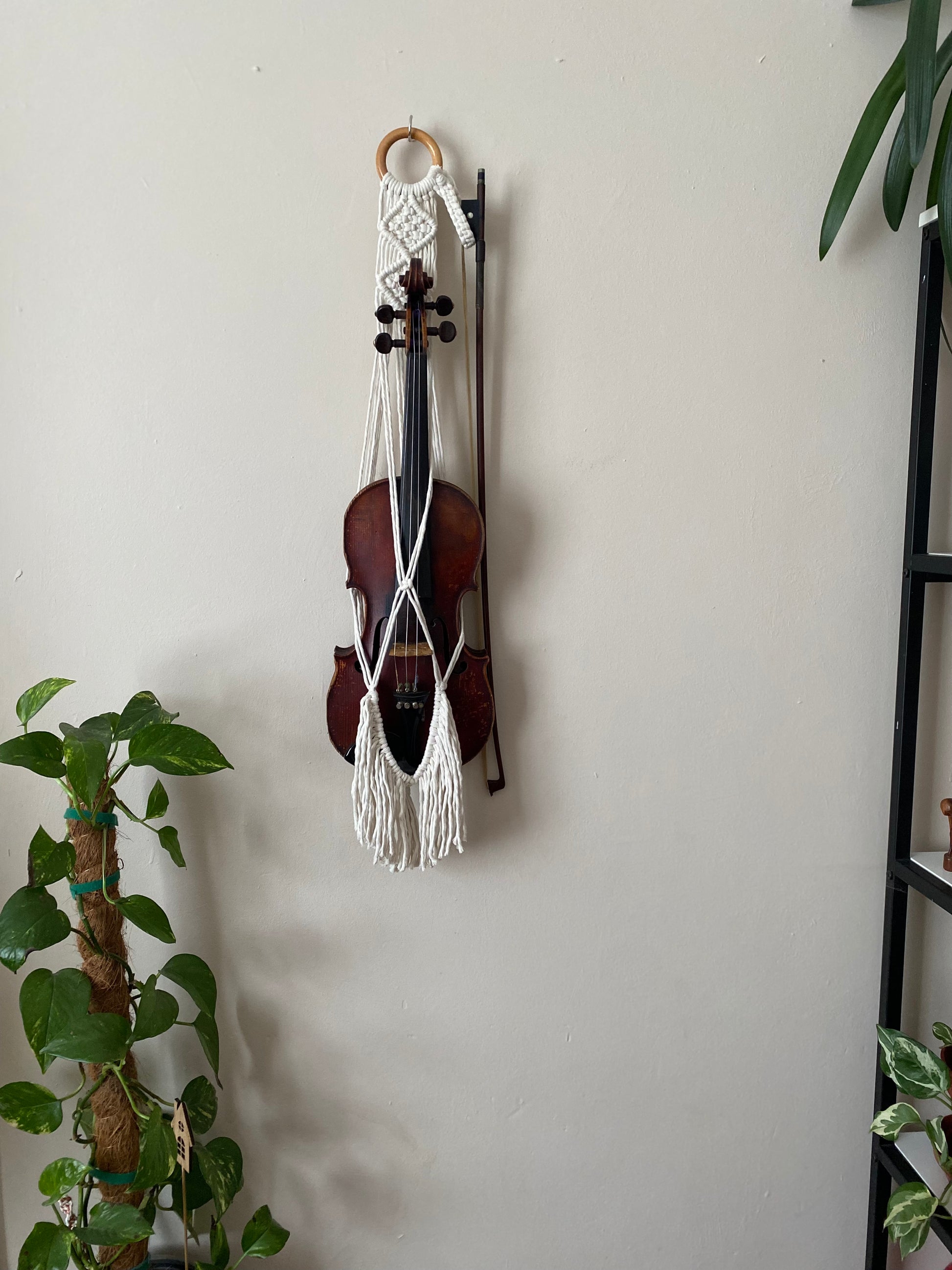 A dark wooden violin hanging in a macrame wall hanger, made with natural off white coloured recycled cotton with tasseled fringe on a wooden ring.