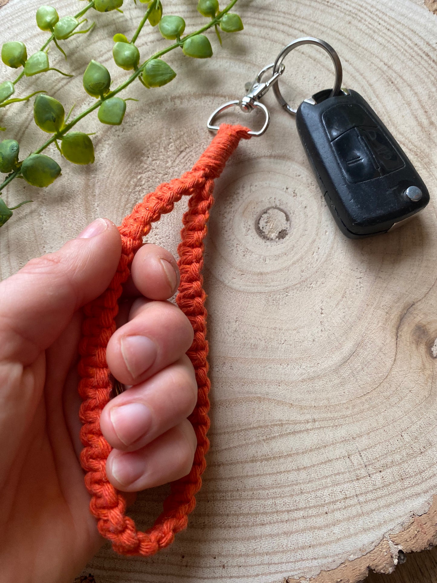 A hand holding a macrame wristlet keychain with metal clasp, made with orange recycled cotton. Shown attached to a single car key.