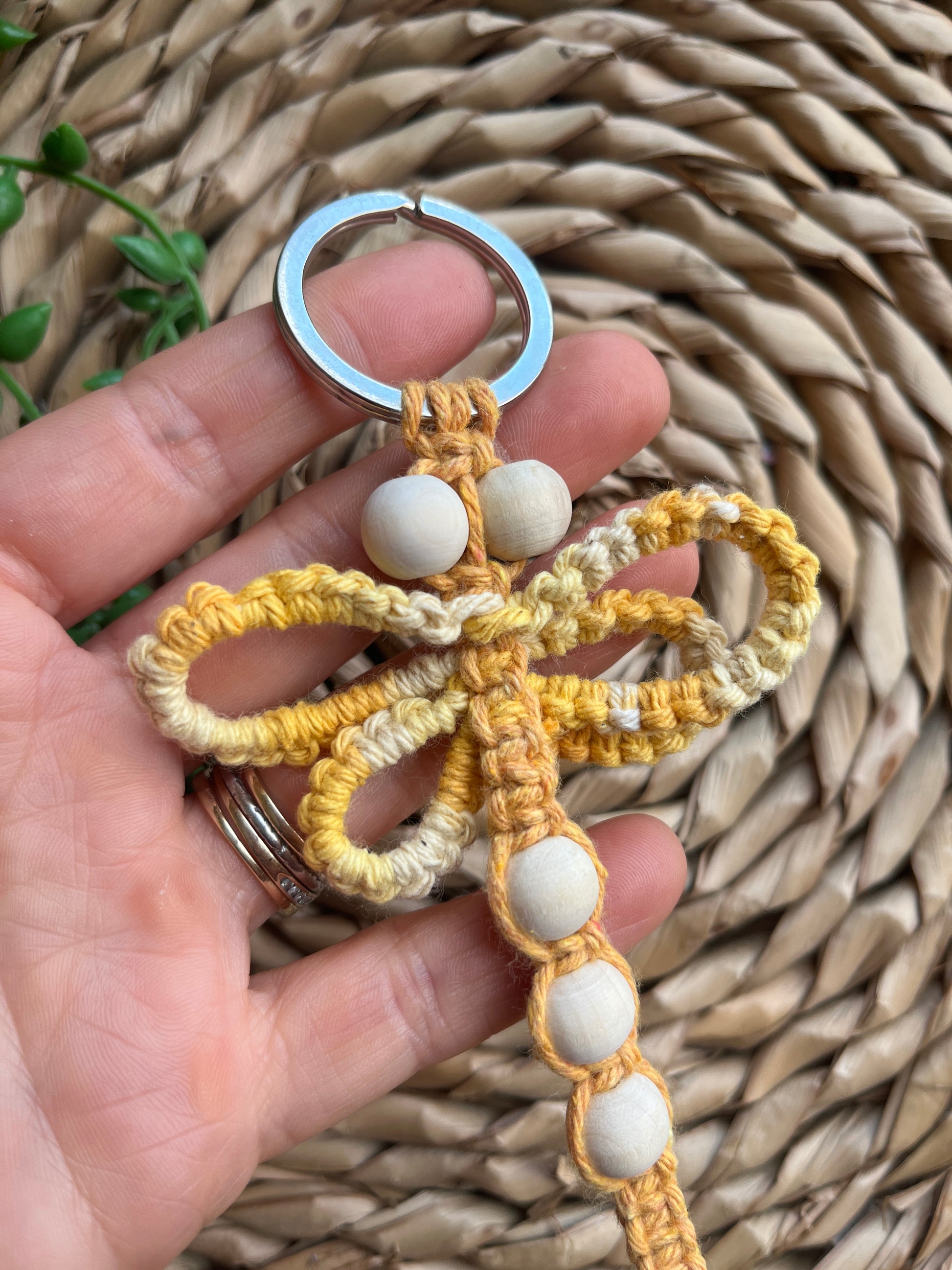 A hand holding a handmade macrame dragonfly keyring or bag charm, made using shades of yellow recycled cotton, on a metal split ring, with small wooden beads.