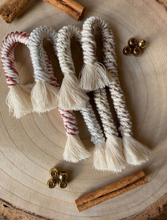 A set of 4 handmade macramé metallic candy cane decorations in silver, gold, copper and white yarn, displayed on a wooden surface with cinnamon sticks and decorative beads.