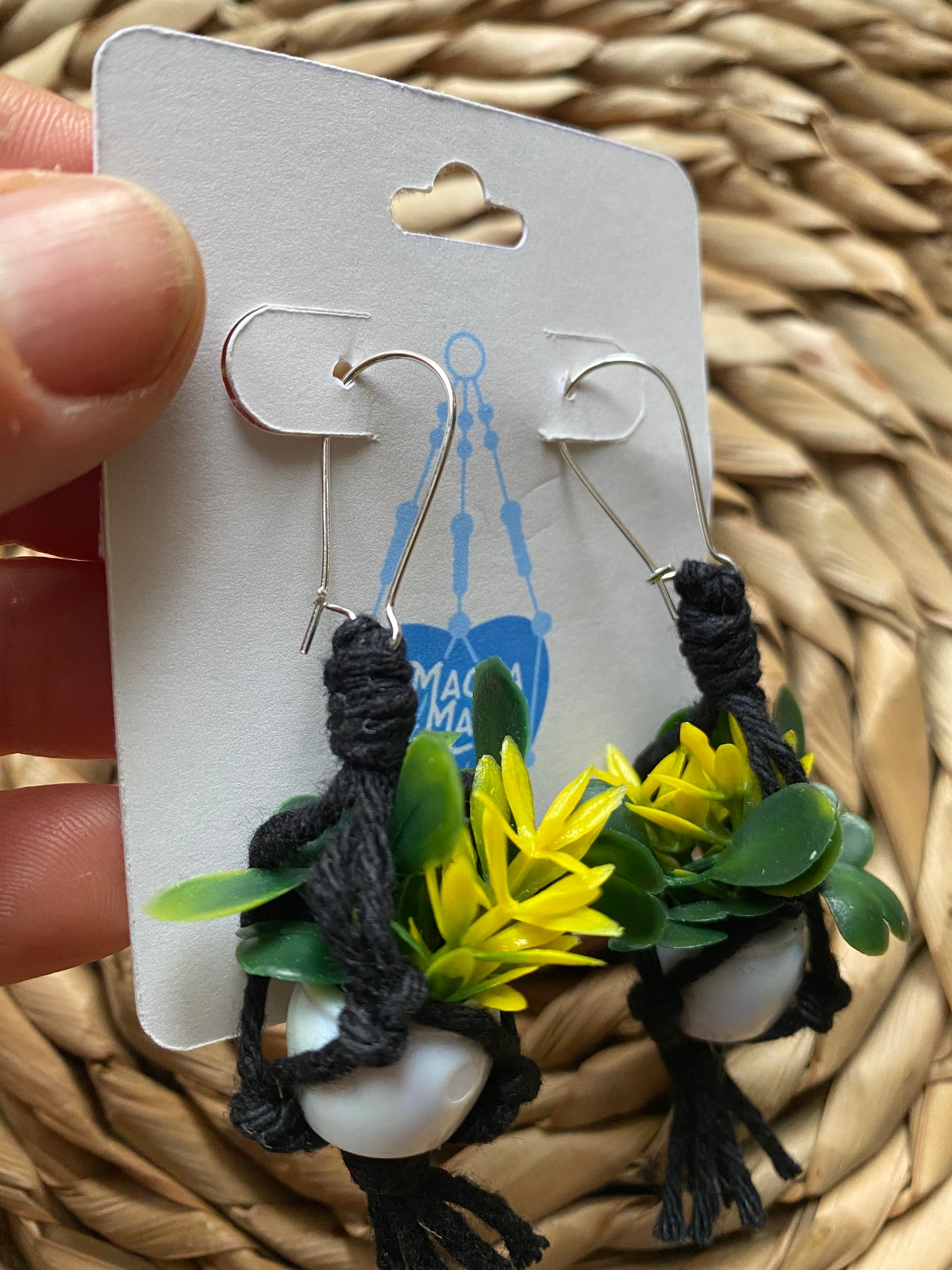 A close up of a hand holding pair of mini macrame plant hanger earrings made with black yarn and small artificial plants, with kidney wire hooks, on a white backing card.