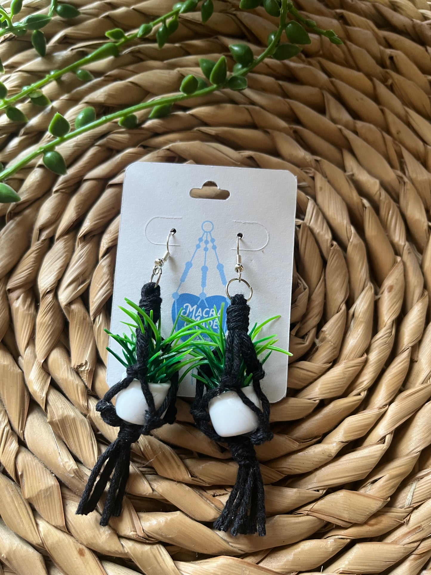 A pair of mini macrame plant hanger earrings made with black yarn and small artificial plants, with silver hooks, on a white backing card, on a wicker surface.