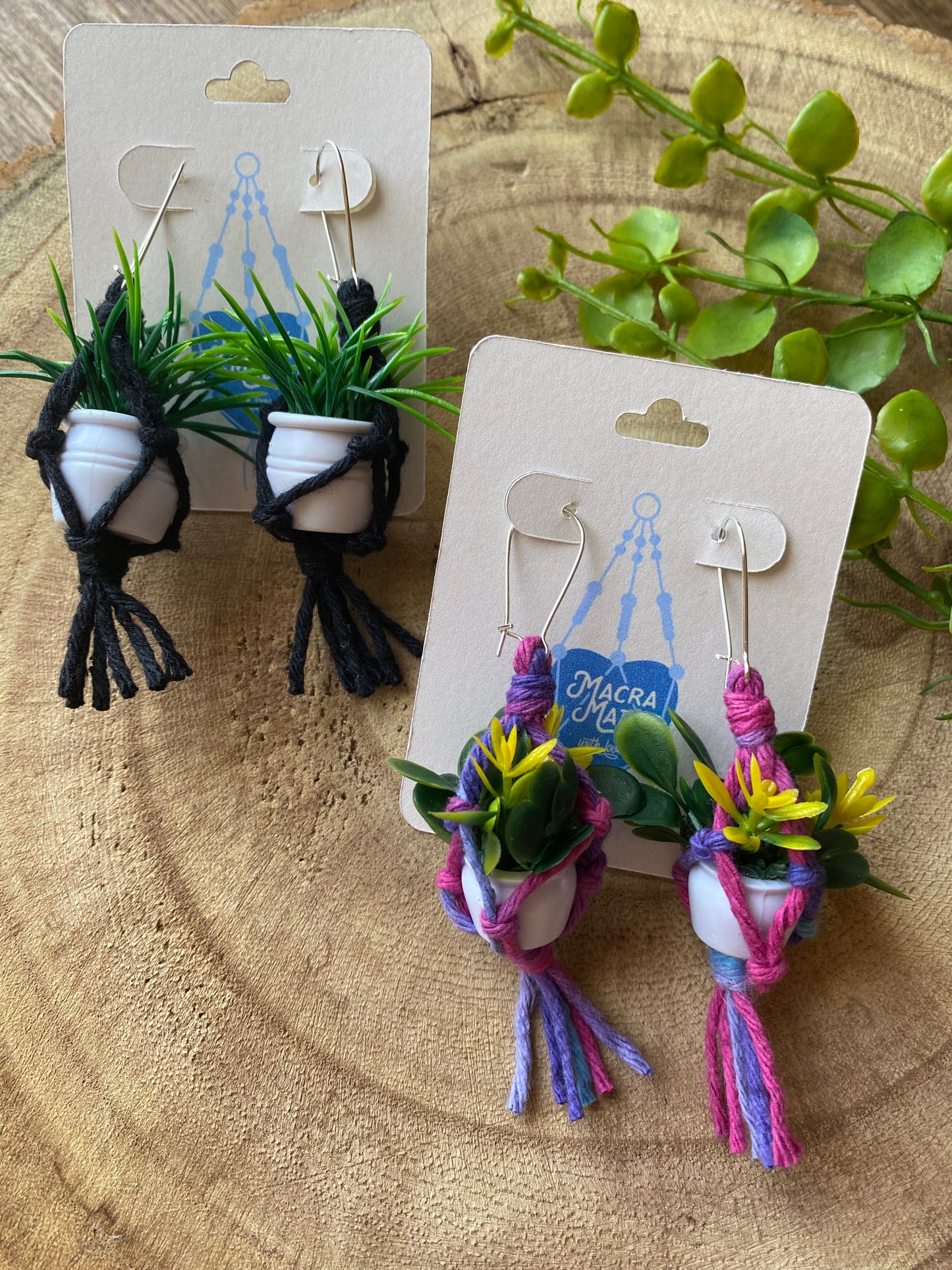 Two pairs of mini macrame plant hanger earrings, one pair made with black yarn and the other pair made with purple yarn, both with small artificial plants and silver kidney wires, on a white backing card, on a wooden surface.