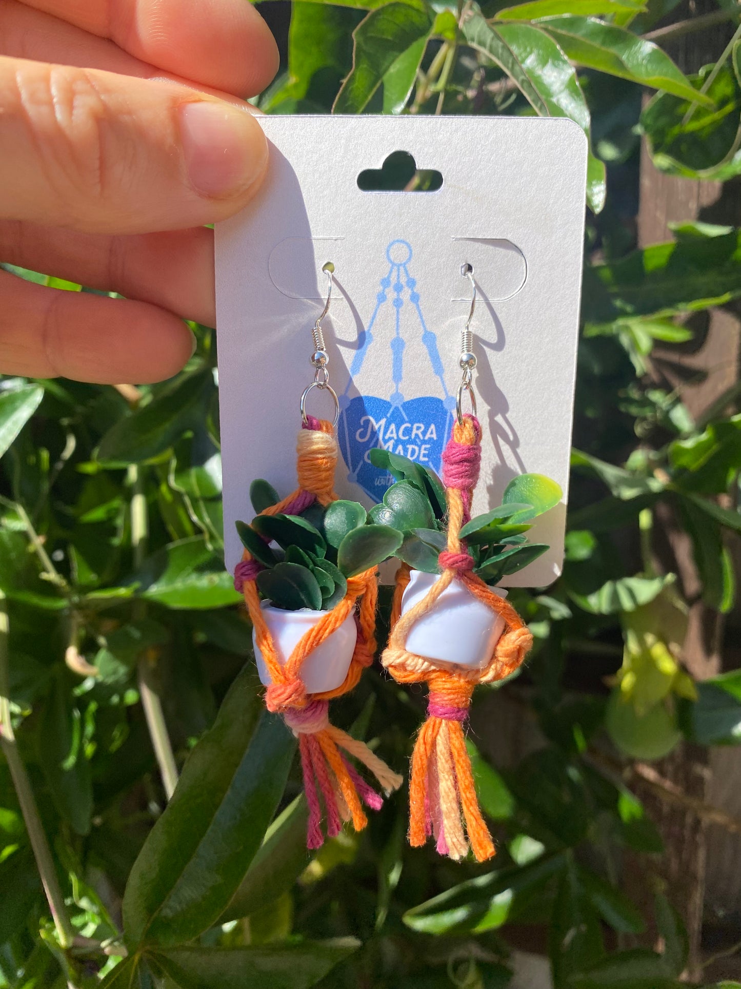 A pair of mini macrame plant hanger earrings made with orange ombre yarn and small artificial plants, with silver hooks, on a white backing card, held outside with lush green plants behind.