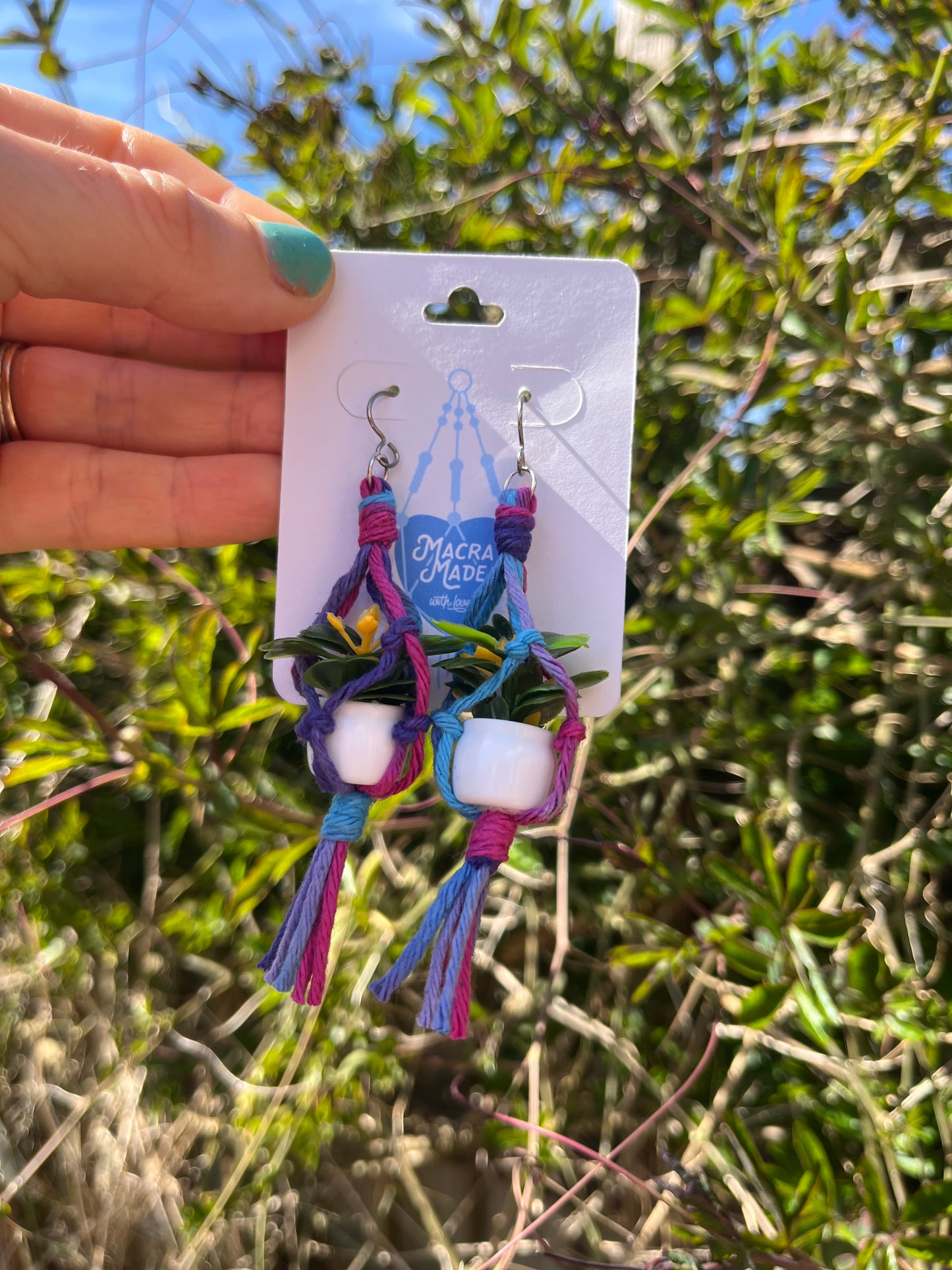 A hand holding a pair of mini macrame plant hanger earrings made with purple yarn and small artificial plants, with silver hooks, on a white backing card, outside with green plants behind.