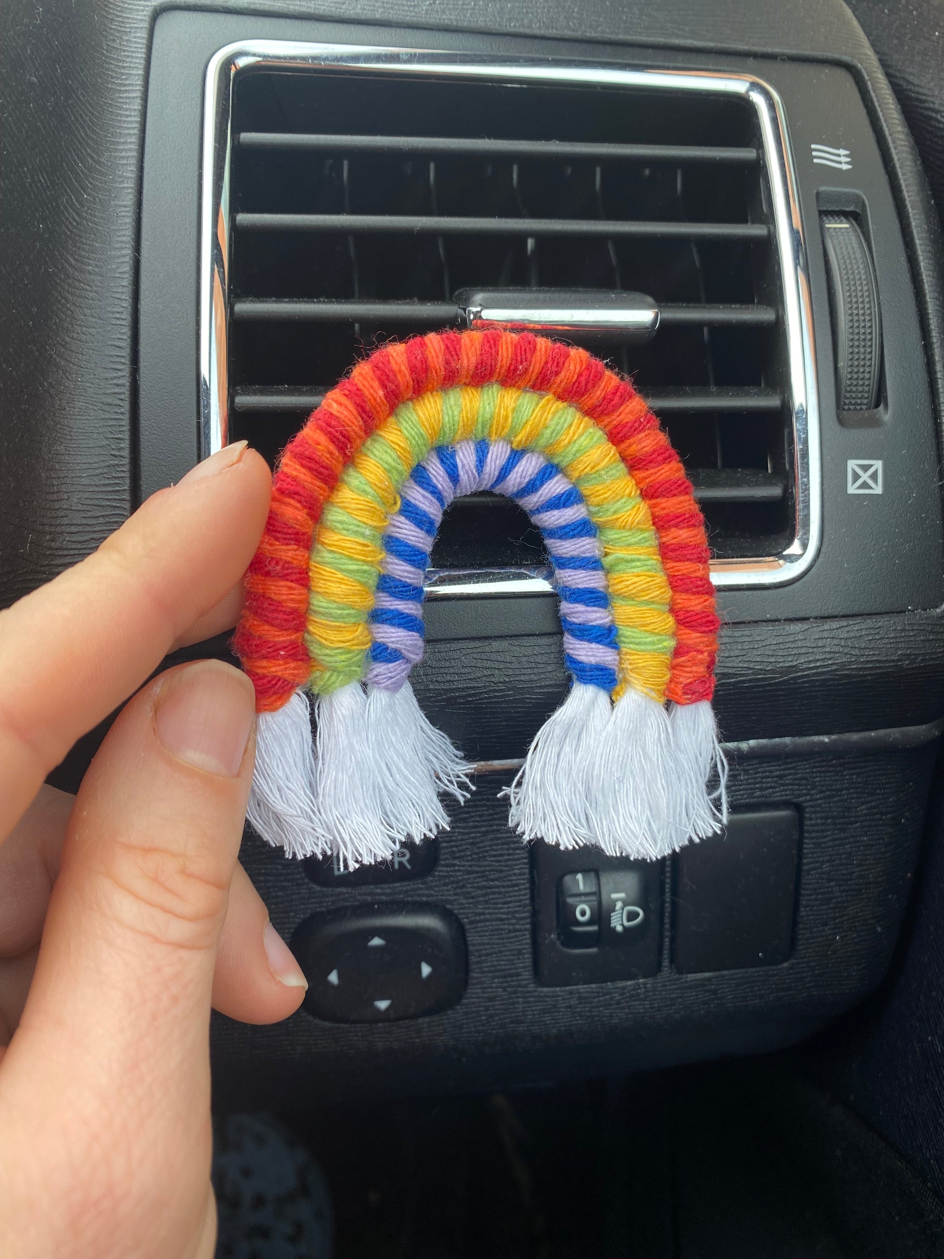 A hand touching a mini macrame rainbow, attached to the car air vent on a black car interior.