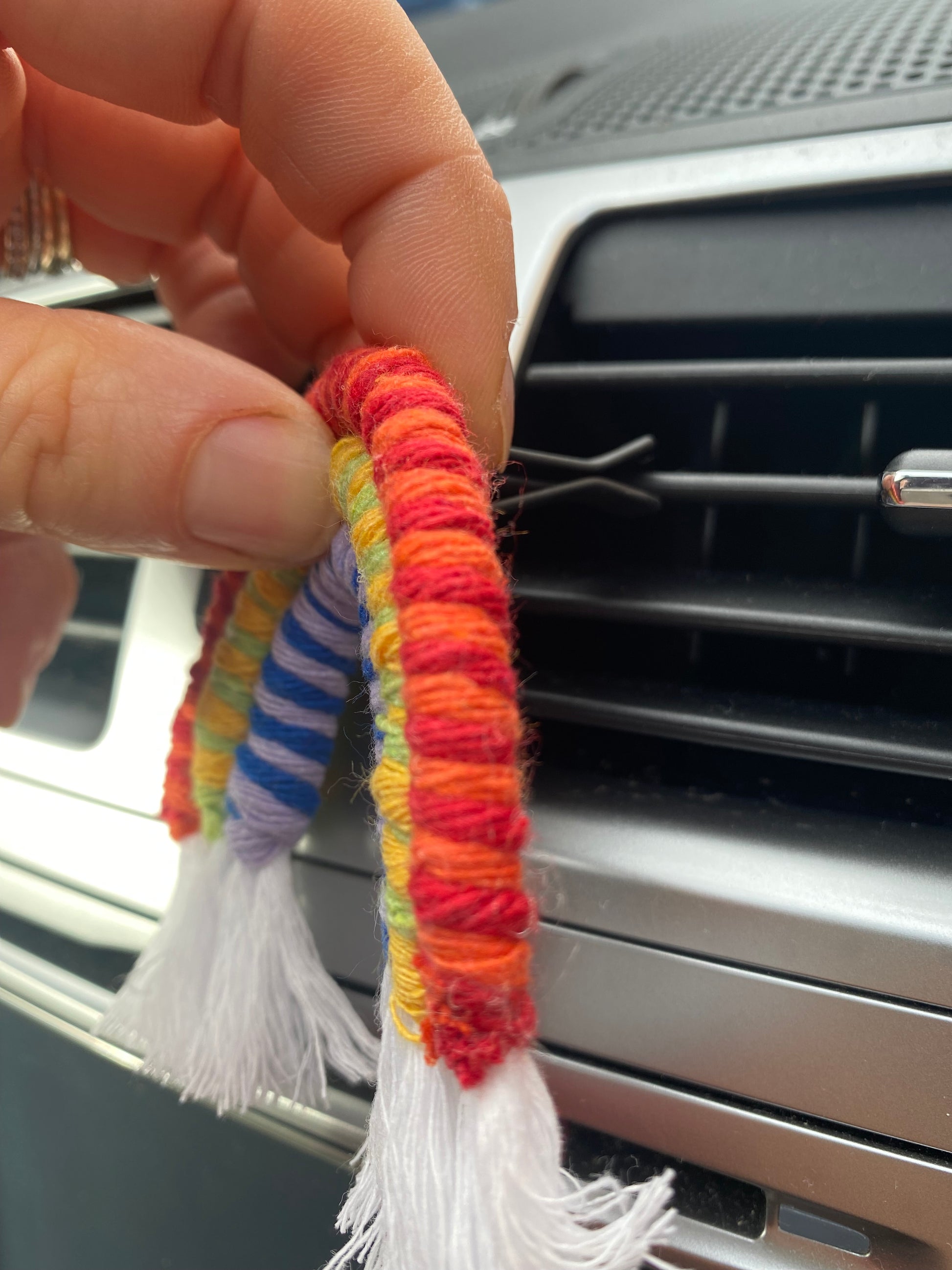 A hand touching a mini macrame rainbow, attached to the car air vent on a black car interior.
