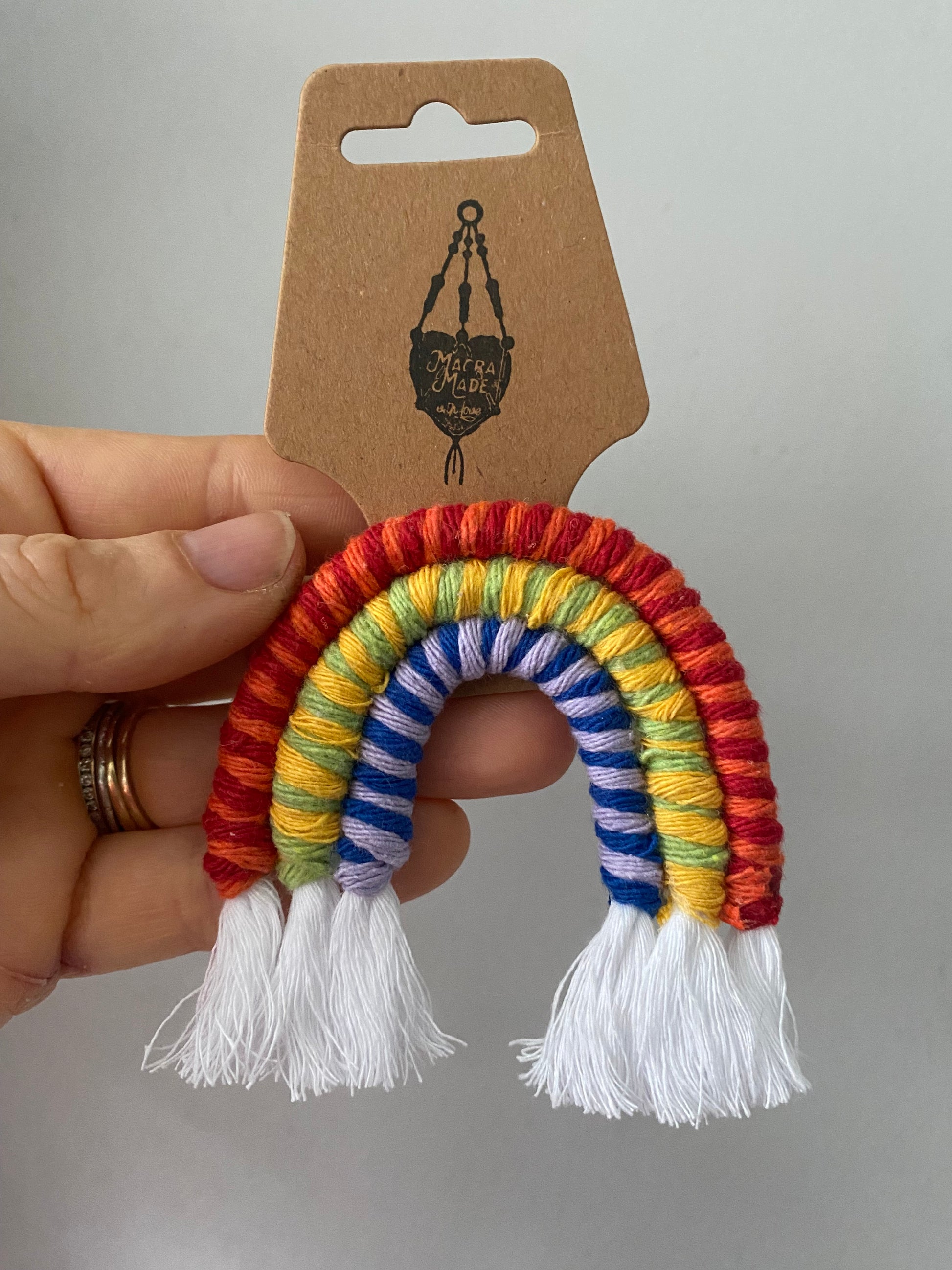 A hand holding a mini macrame rainbow, attached to a cardboard swing tag.
